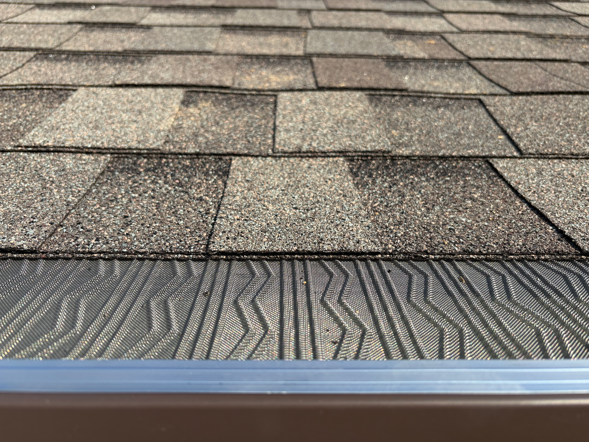 A textured, gray gutter guard installed under the edge of brown asphalt roofing shingles.
