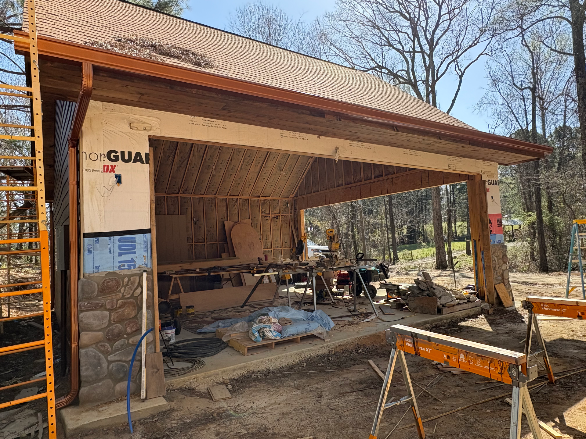 An unfinished structure with stone base and metal roofing under construction, surrounded by trees.