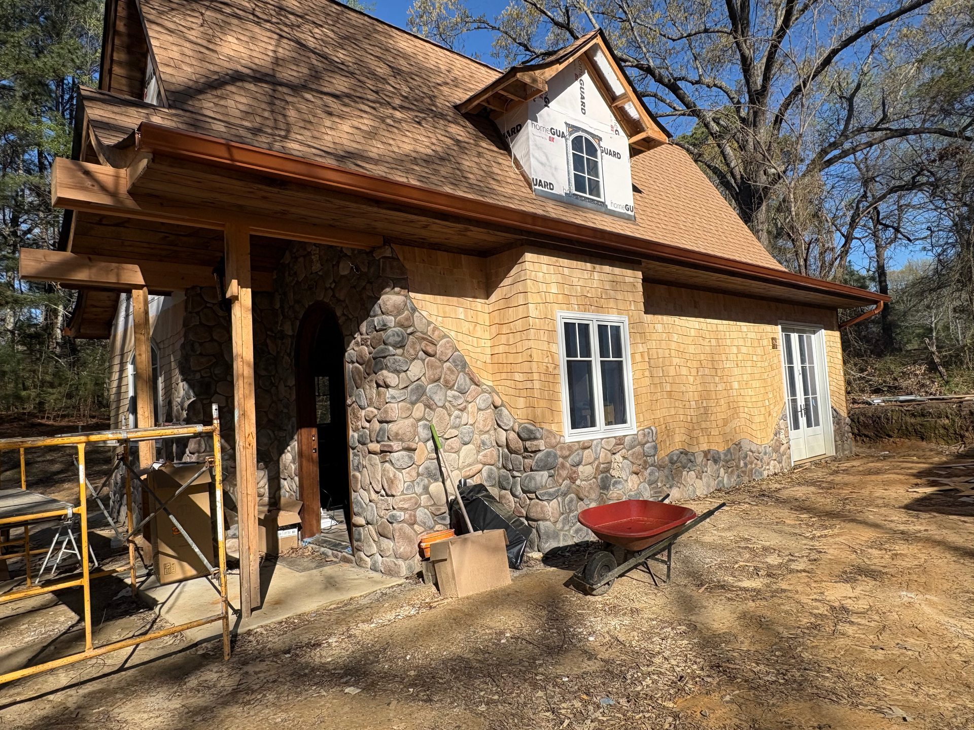 A rustic house under construction, featuring stone siding on the lower half, brown wooden walls, and a gabled roof.