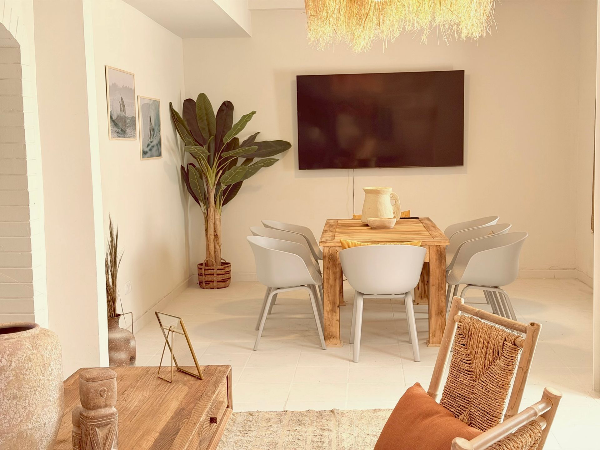 Comedor minimalista blanco con mesa de madera, bancos, planta en maceta y lámpara colgante de mimbre.