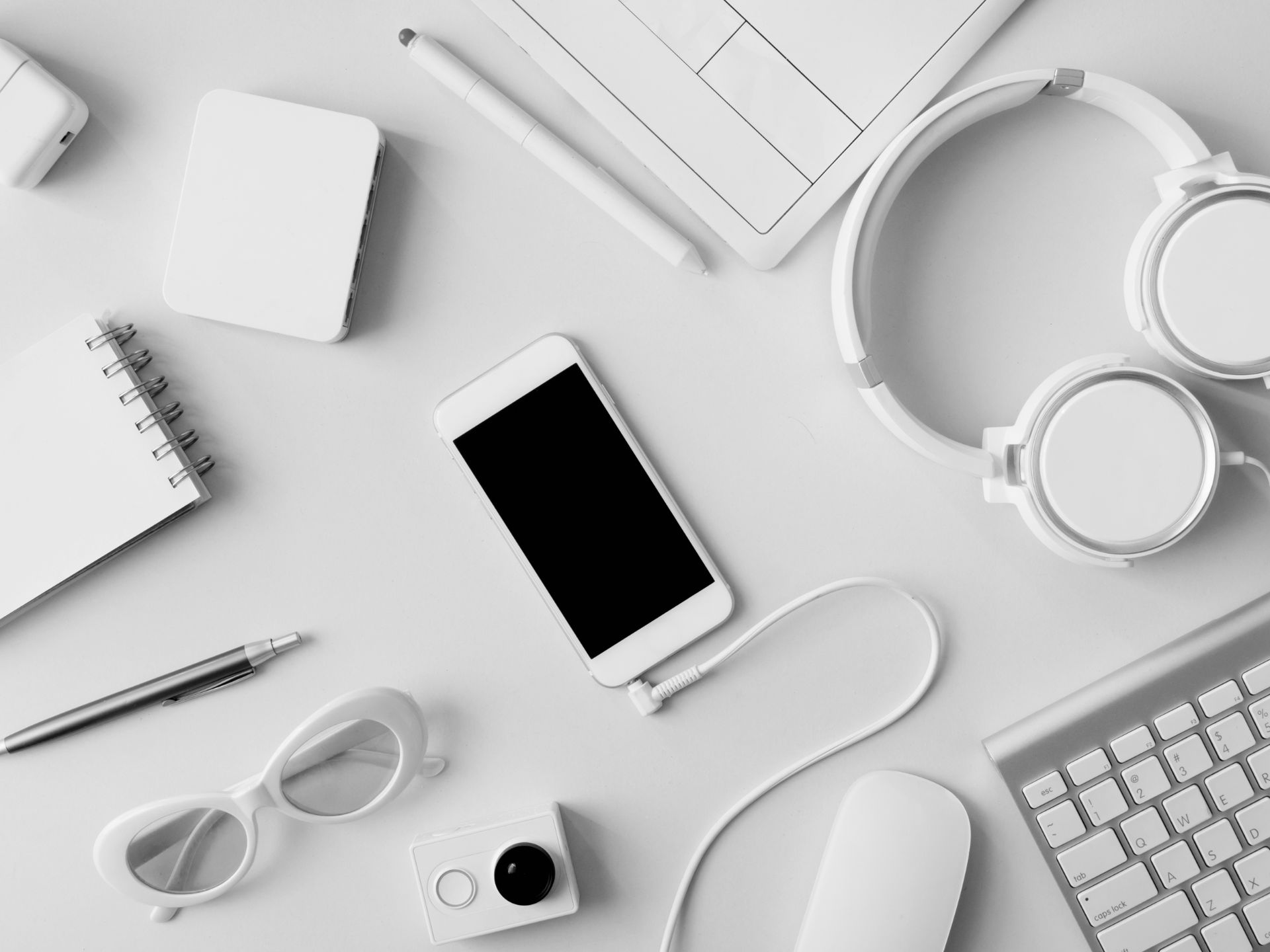 A white desk with a cell phone , headphones , a camera , and a keyboard.