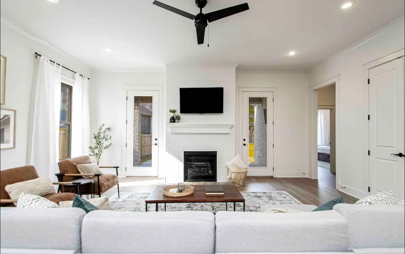 Spacious living room with white walls, a black ceiling fan, and hardwood floors. Brown leather chairs and a white couch sit on a rug.