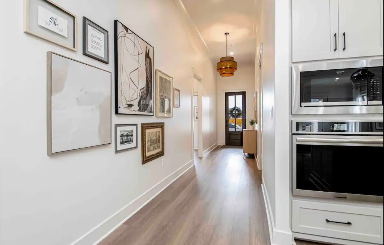 Long hallway with artwork on the left, kitchen cabinetry with appliances on the right, and a door at the end.