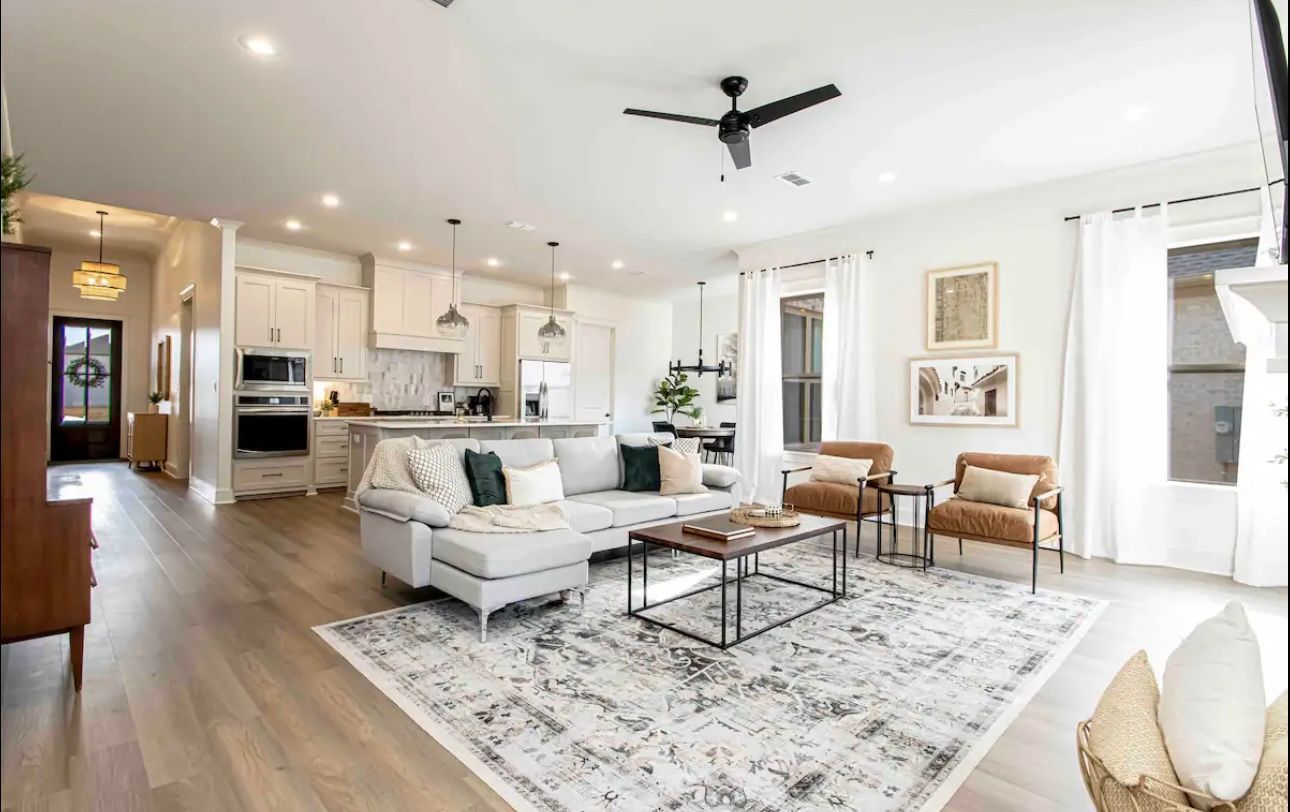 Open-concept living space with kitchen, living room, and dining area. Neutral colors, light wood floors, gray sectional, and a patterned rug.