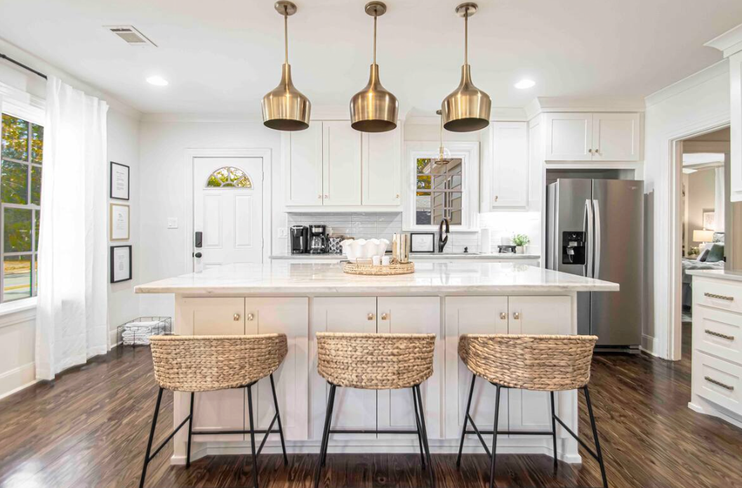 Kitchen with white cabinets, island with woven stools, brass pendant lights, stainless steel appliances, and dark wood floors.