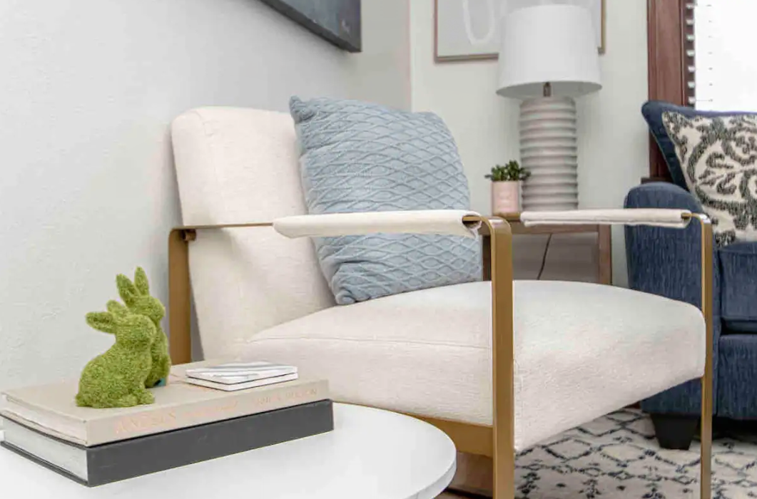 White armchair with gold frame, blue pillow, small side table with books and bunny figurines.