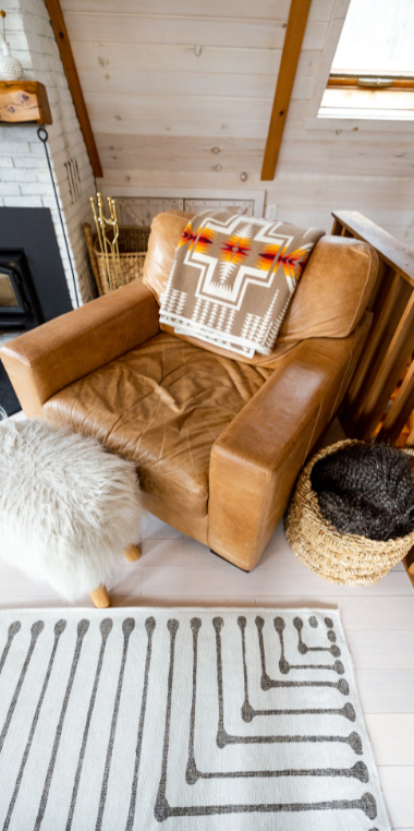 Cozy room with a tan leather armchair, patterned throw pillow, fluffy footstool, woven basket, and geometric rug.