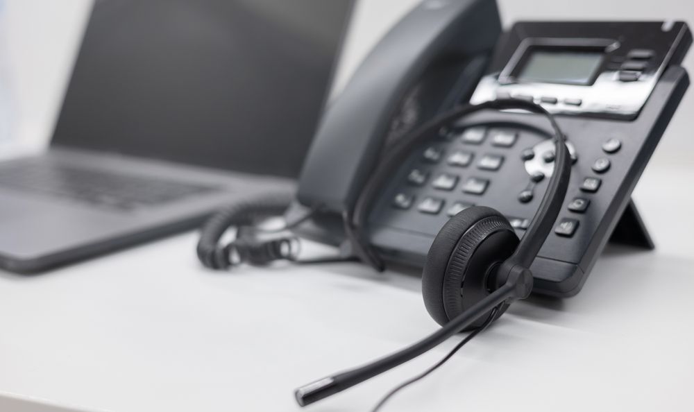 Black office desk phone with headset beside a laptop on a white table.