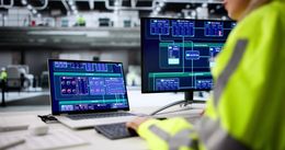 Person in yellow safety vest monitoring industrial control screens at a workstation