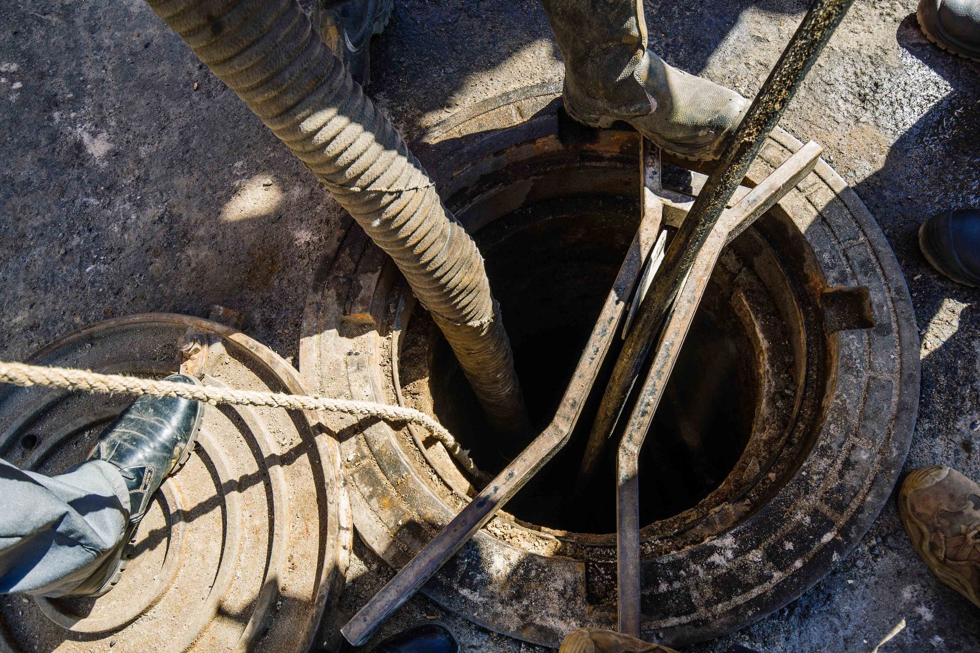 Workers’ boots and hoses around a circular manhole opening in gravel Workers’ boots and hoses around a circular manhole opening in gravel