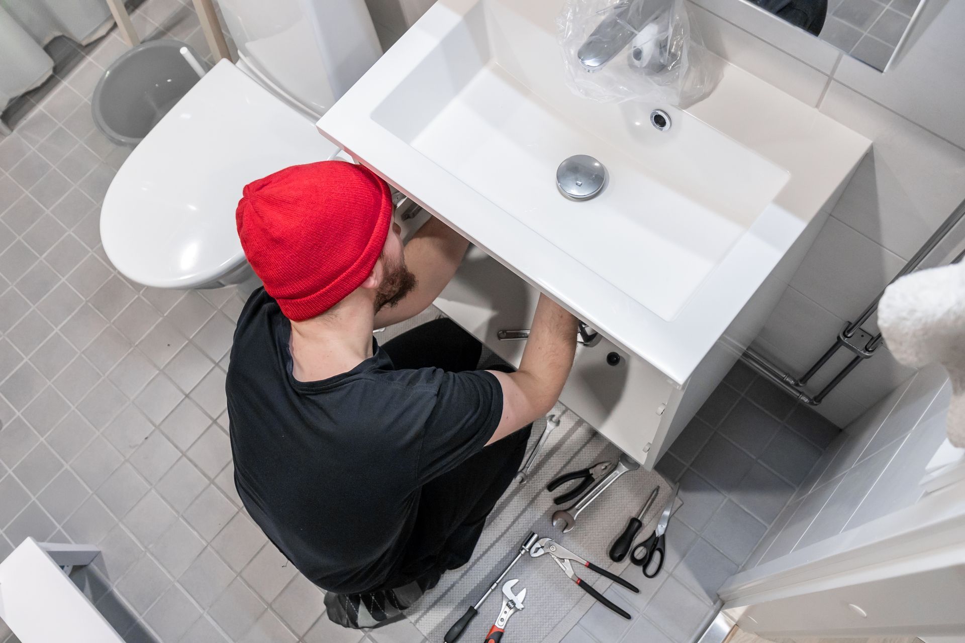 Person in a red beanie fixing plumbing under a bathroom sink with tools on the floor