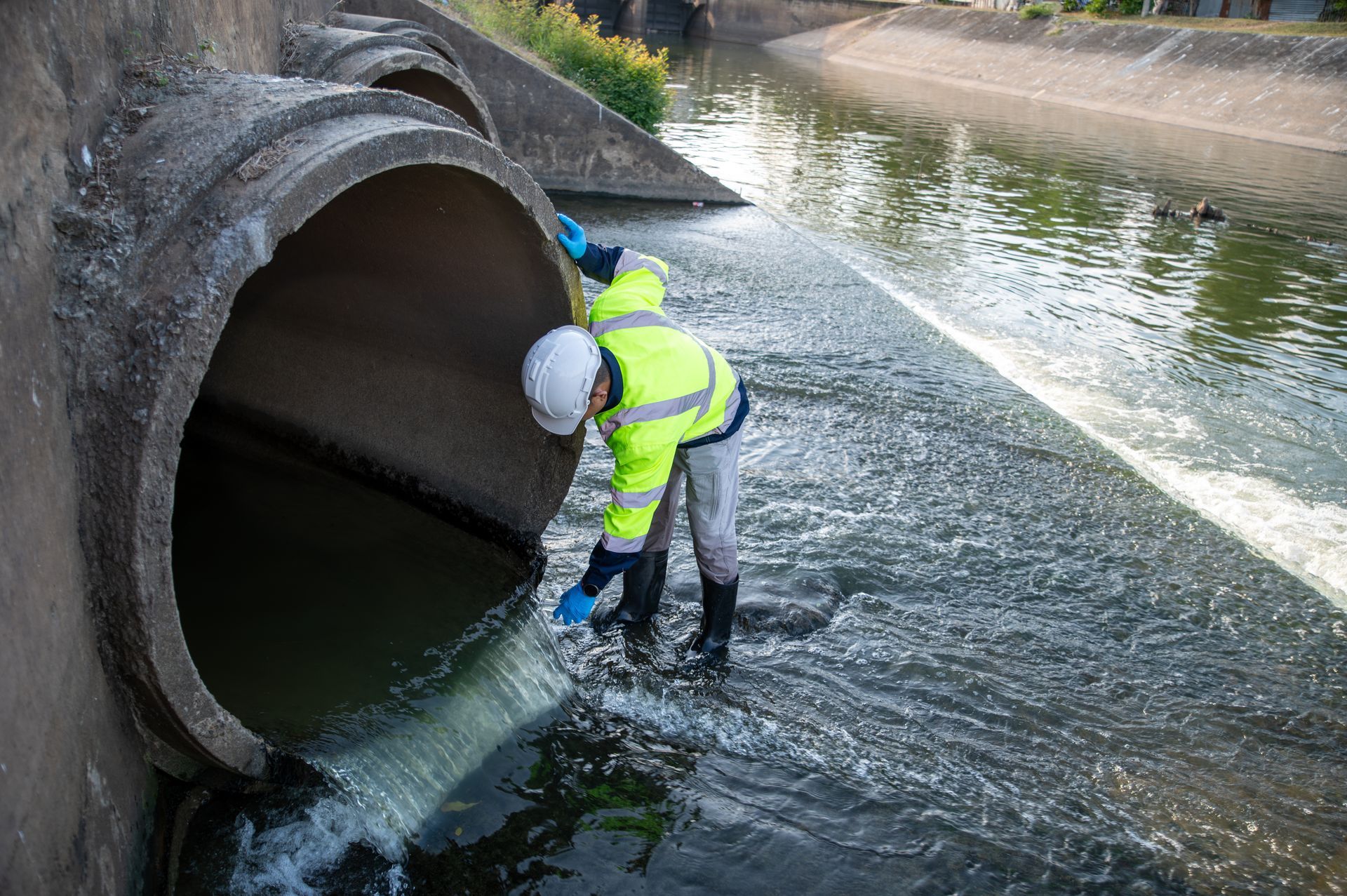 Worker in high-visibility gear inspecting water flowing from a large concrete pipe into a canal Worker in high-visibility gear inspecting water flowing from a large concrete pipe into a canal