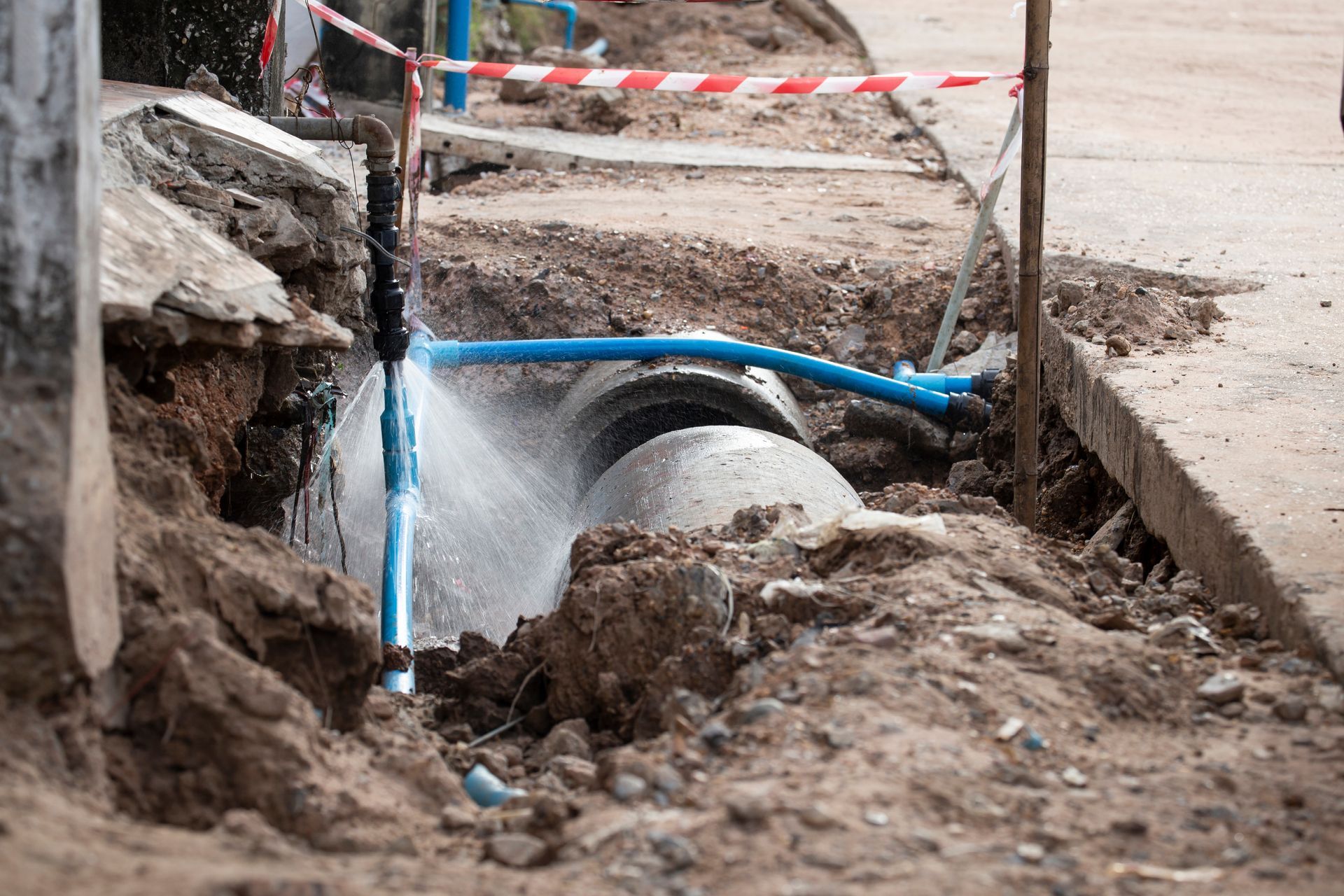 Broken water pipe spraying in a muddy roadside trench, with blue piping and caution tape nearby Broken water pipe spraying in a muddy roadside trench, with blue piping and caution tape nearby