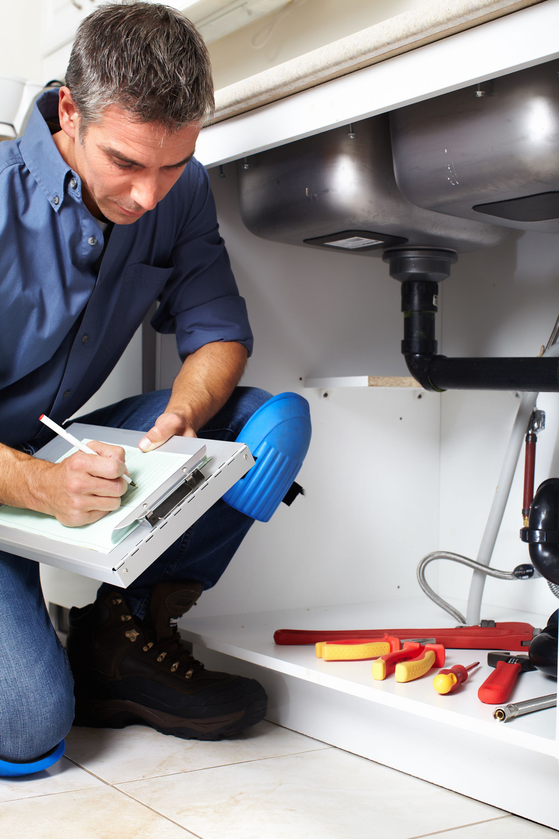 Plumber inspecting under-sink pipes with a clipboard and tools in a cabinet.