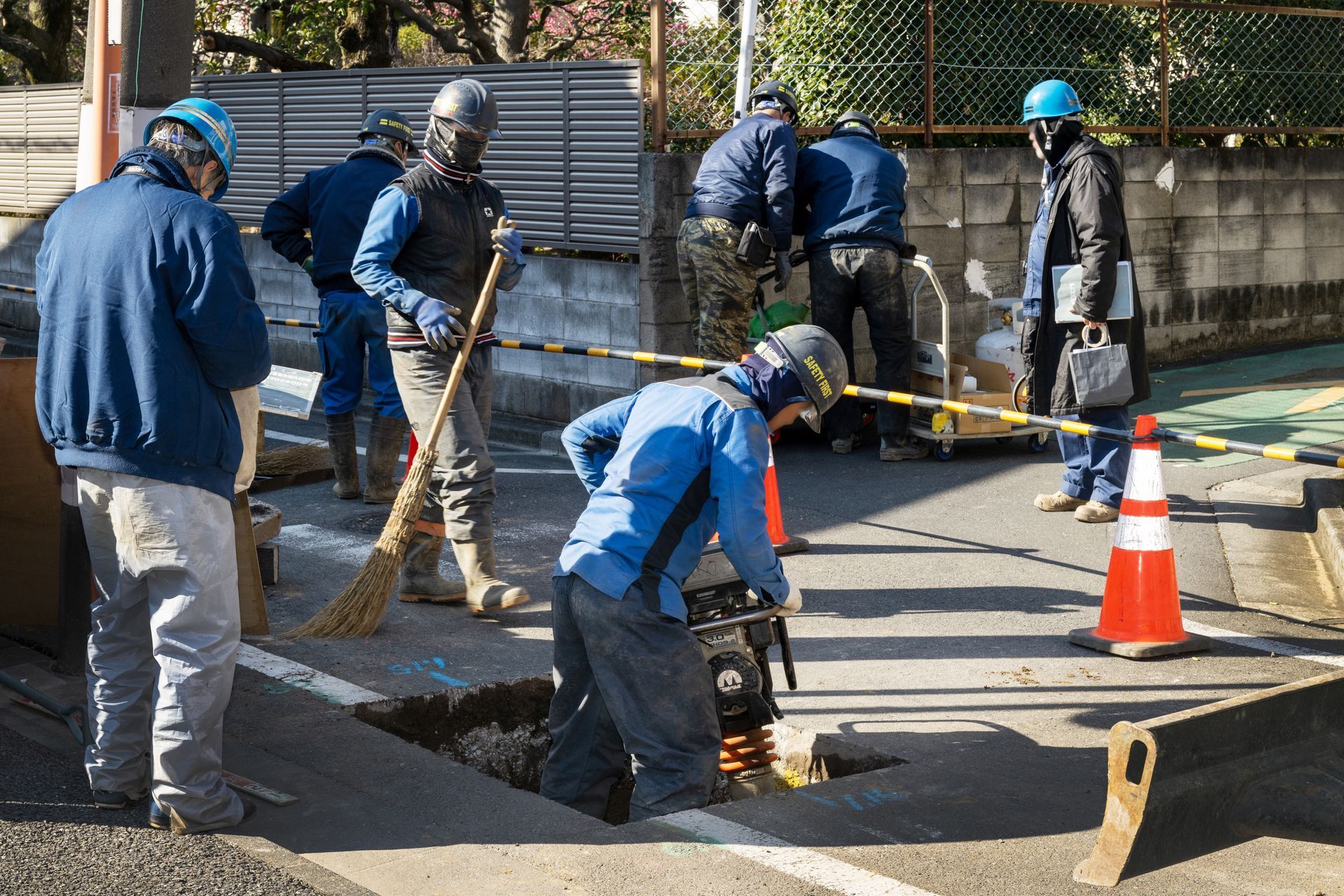 Workers in blue uniforms repair a street, shoveling and paving beside an orange traffic cone. Workers in blue uniforms repair a street, shoveling and paving beside an orange traffic cone.