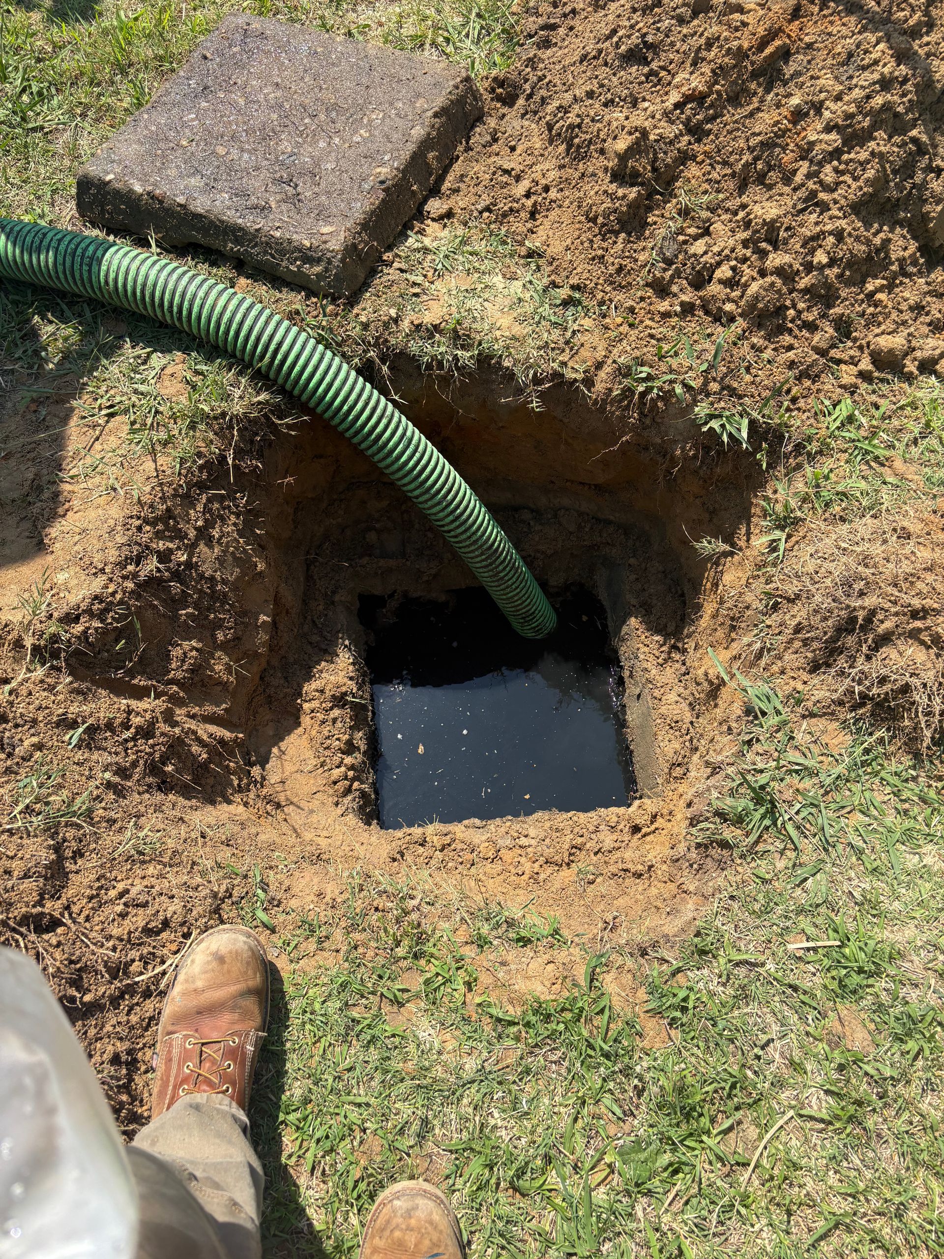 Green hose feeding into a square ground hole beside a removed paver and dug-up dirt patch