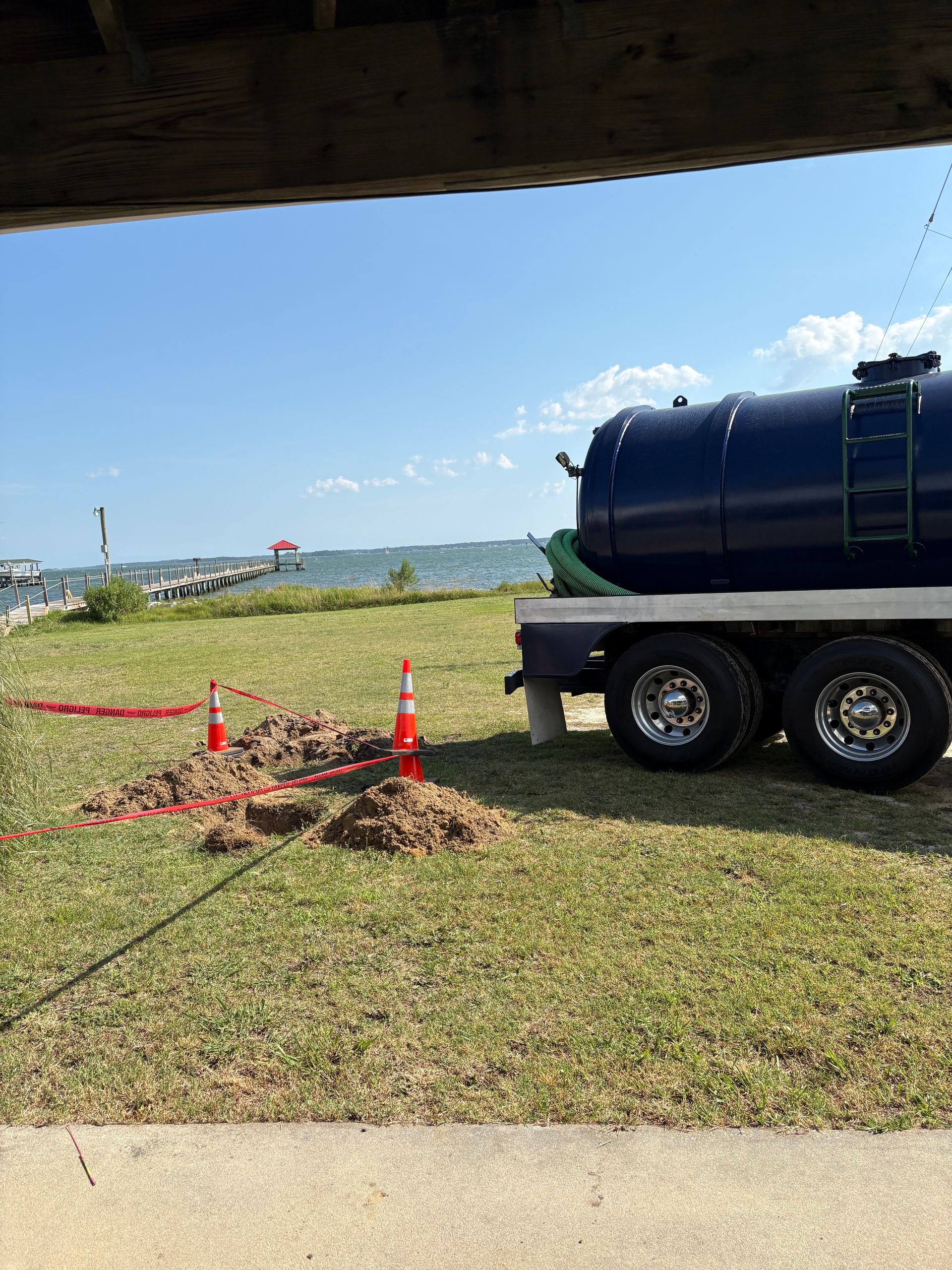 Blue tanker truck beside a small roadwork area with orange cones and tape on a grassy roadside by the water