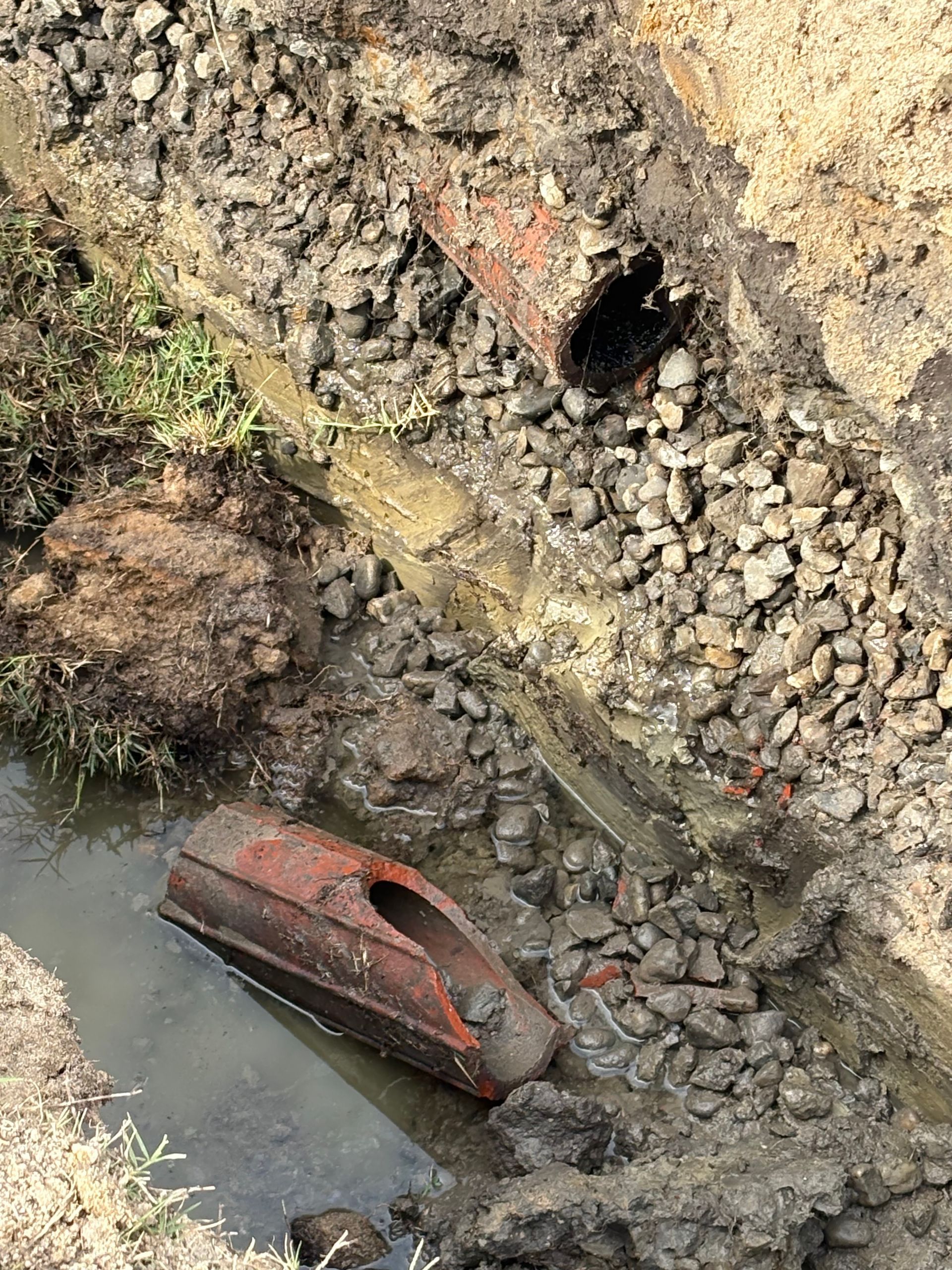 Broken red pipes in a muddy ditch beside standing water, with gravel and dirt embankment.