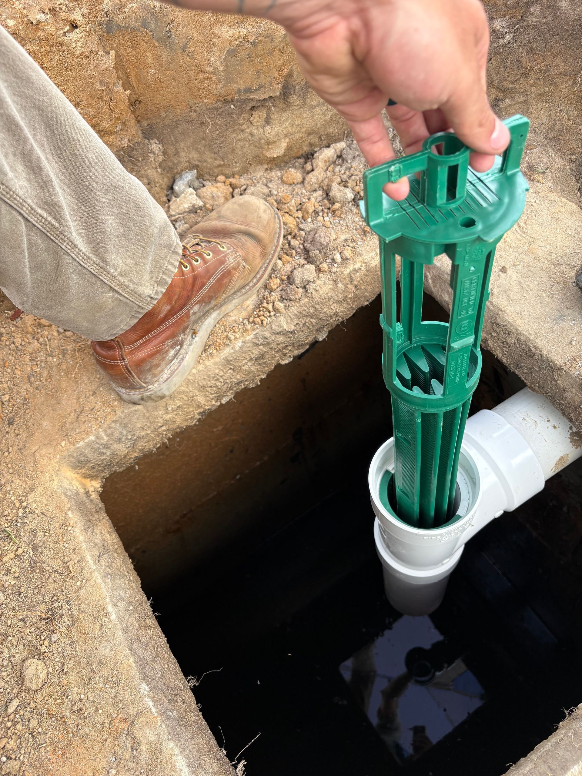 Hand placing a green plastic tool into a white PVC pipe opening beside a dark pit Hand placing a green plastic tool into a white PVC pipe opening beside a dark pit