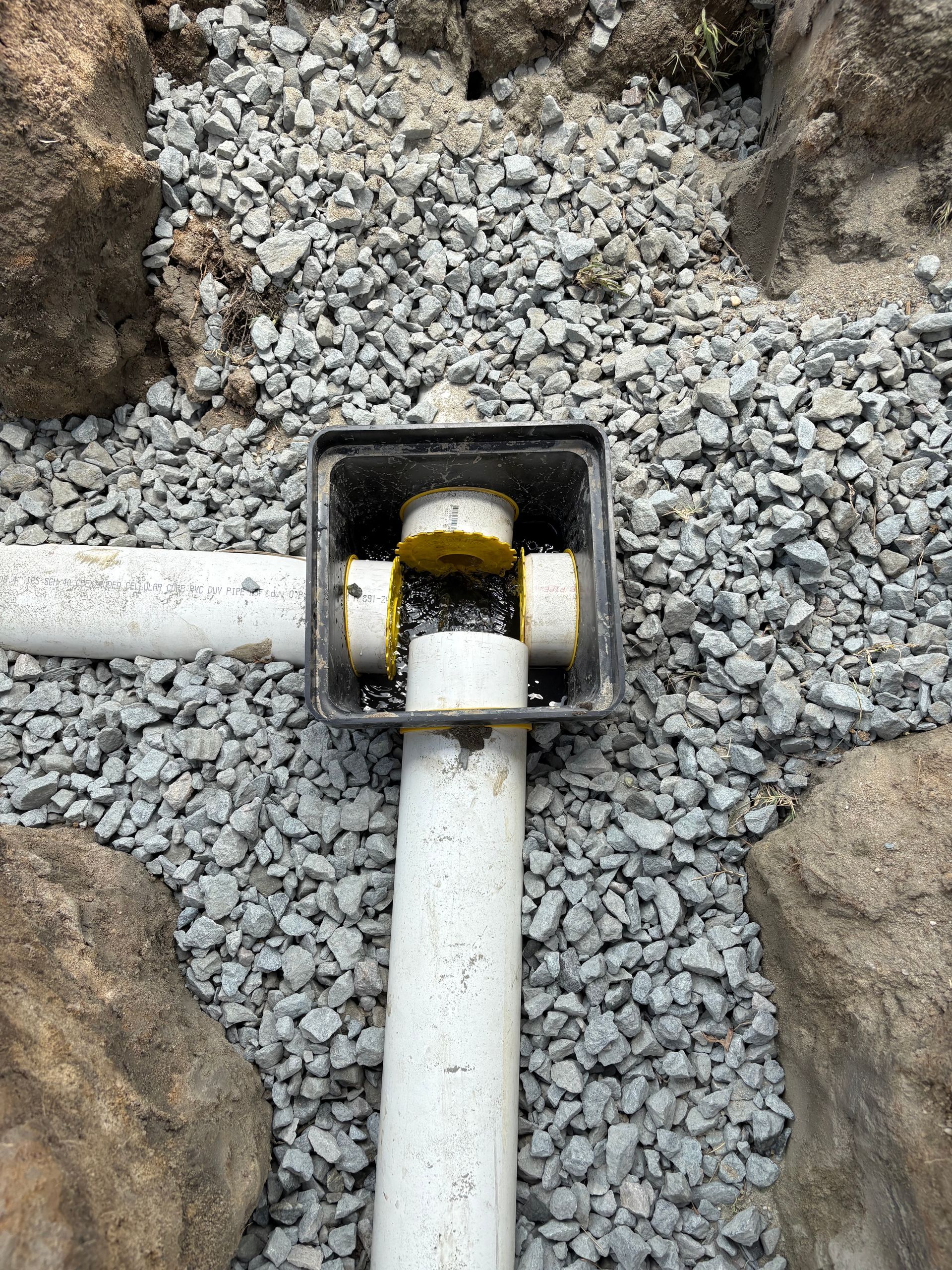 White pipes join in a black square fitting amid gray gravel and rocks. White pipes join in a black square fitting amid gray gravel and rocks.