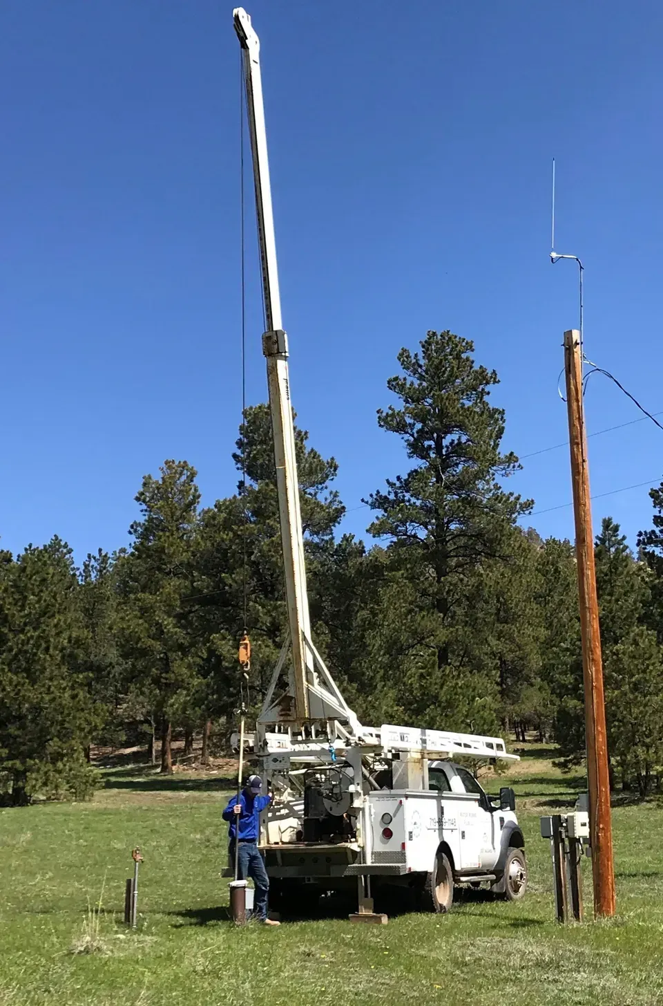 Utility truck with a raised boom beside a wooden pole in a grassy field under a blue sky.