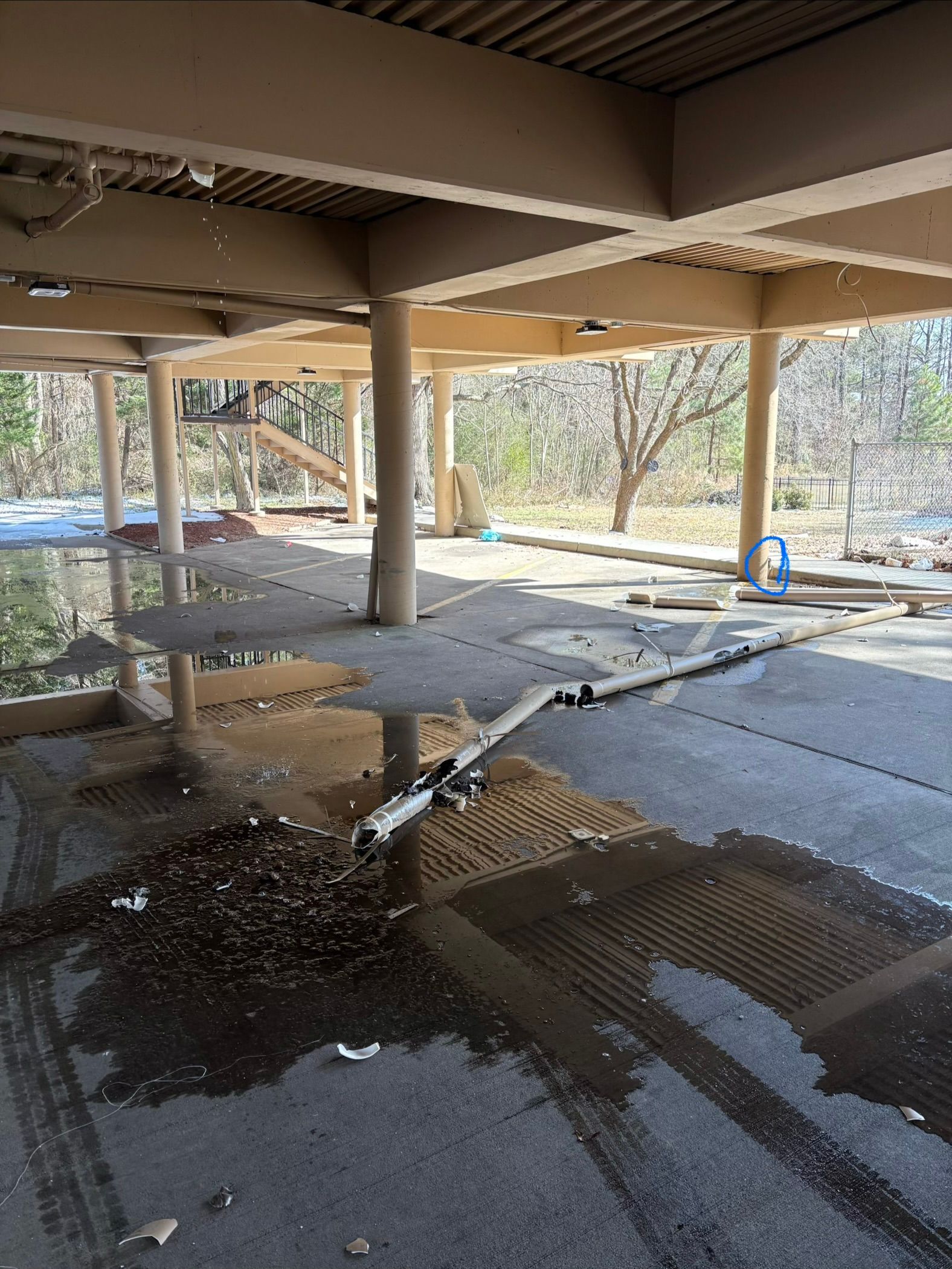Flooded concrete under a shaded structure with puddles reflecting columns and trees beyond.