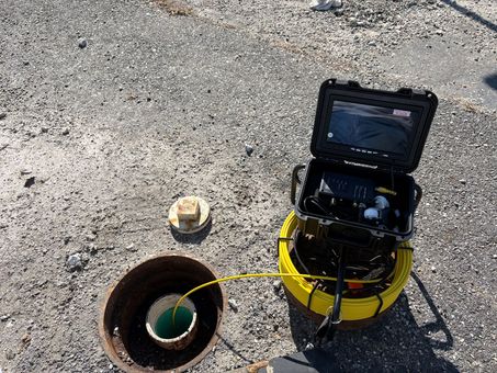 Inspection camera setup beside an open pipe on gravel, with a yellow unit and monitor open.