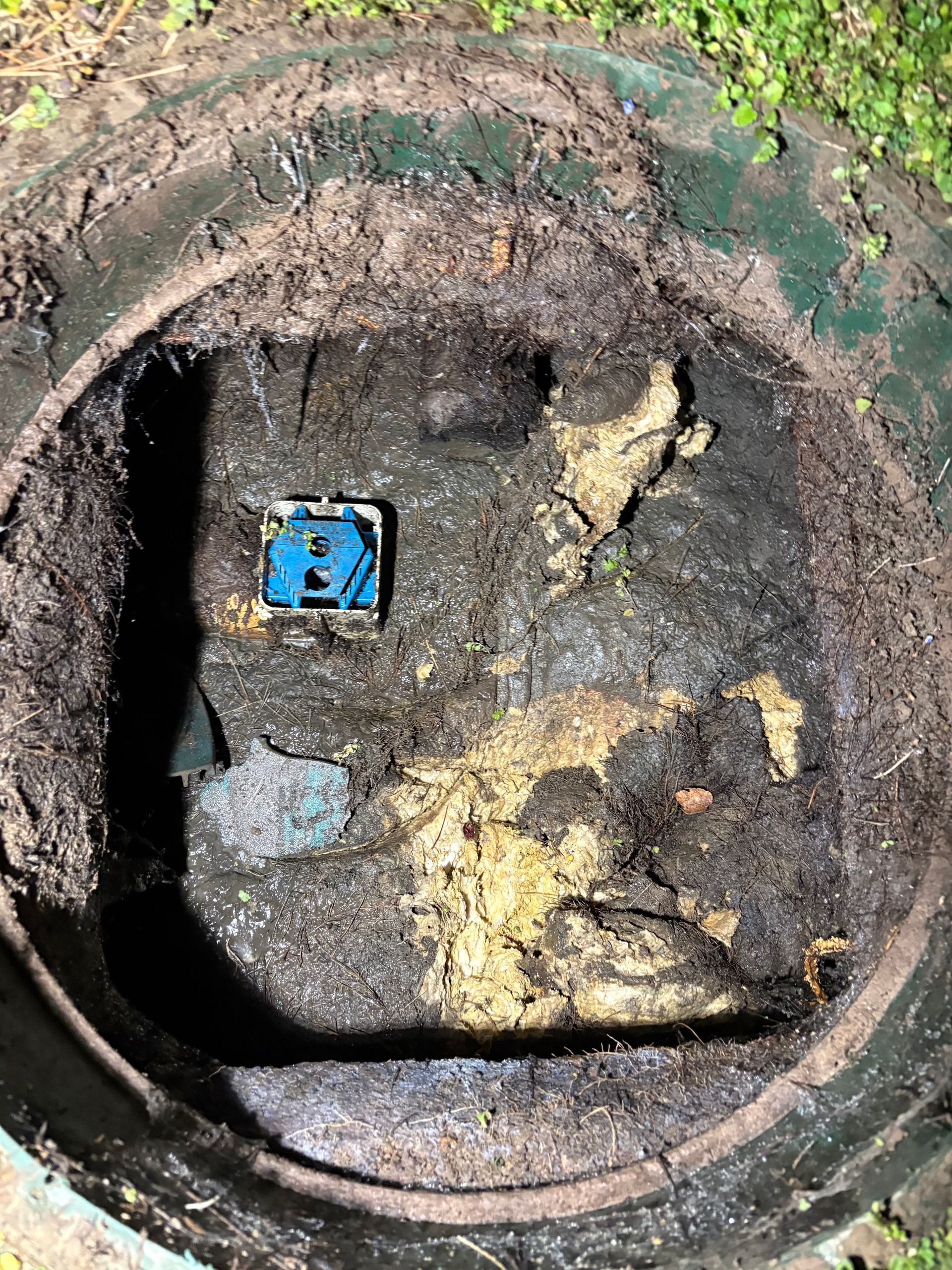 Open round manhole with muddy, debris-filled interior and a small blue valve box inside.