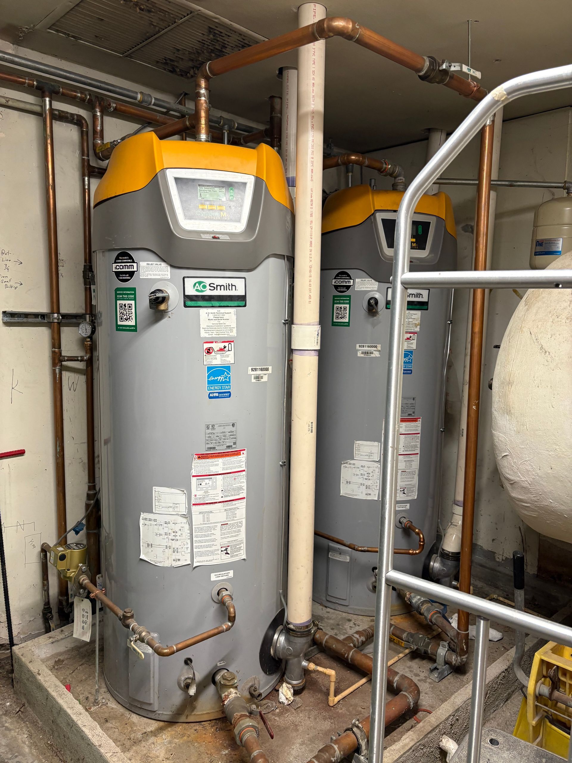 Two gray water heaters with yellow tops in a utility room, surrounded by pipes and a white tank.