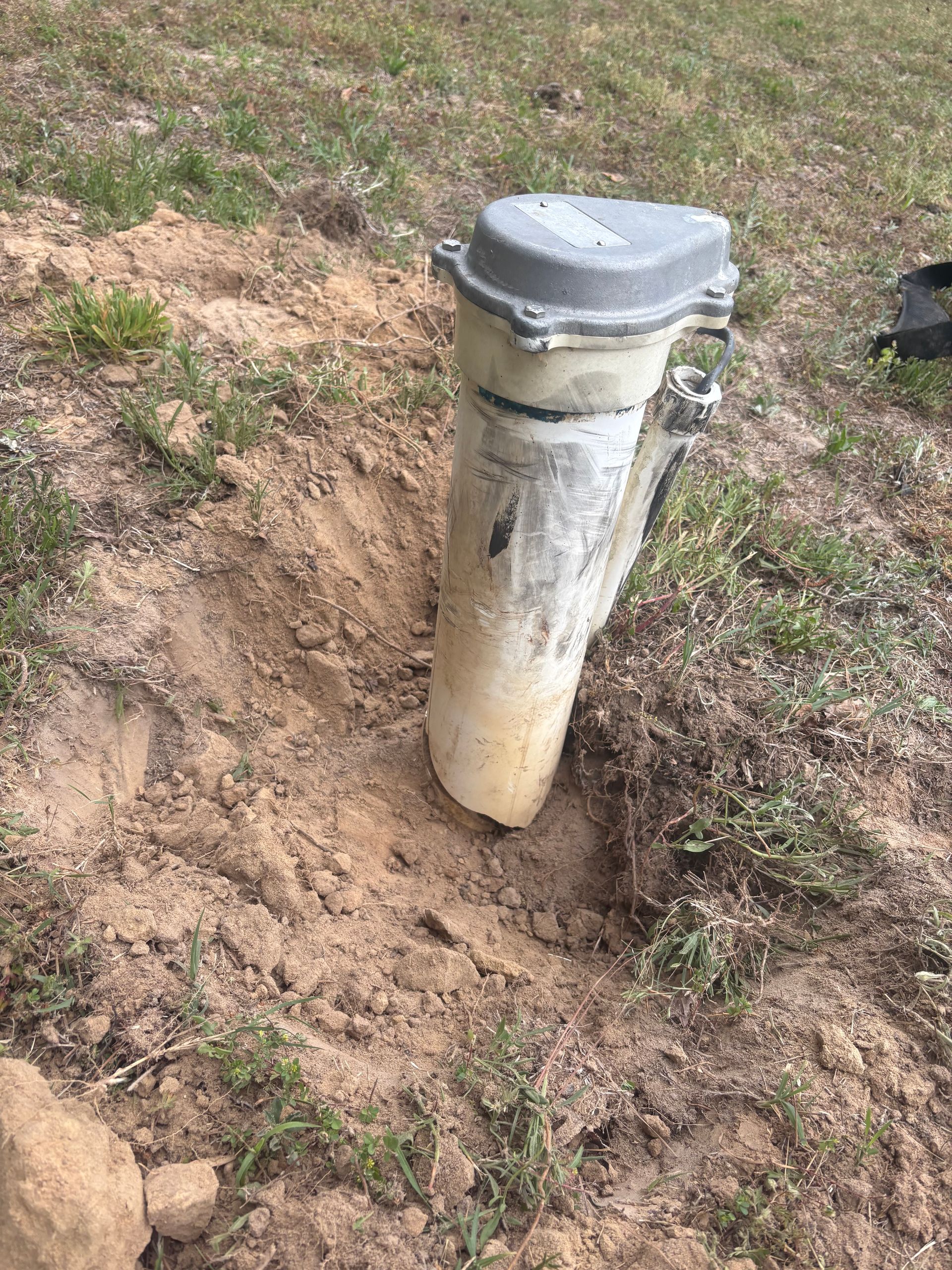 White utility pipe or vent post in a dirt patch beside grass, with a gray cap on top