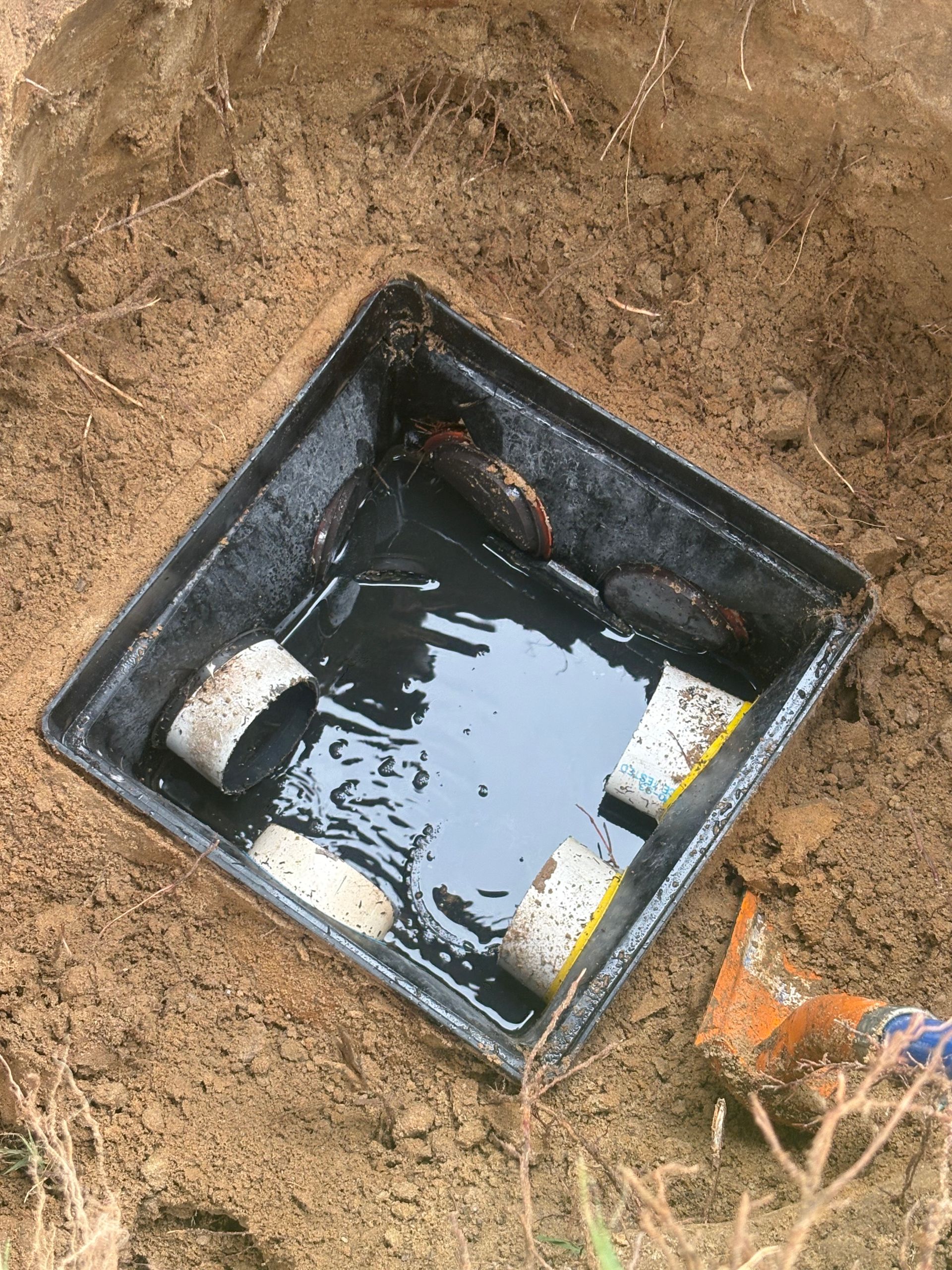 Open square utility box in dirt with water and debris inside, one orange and two white items visible