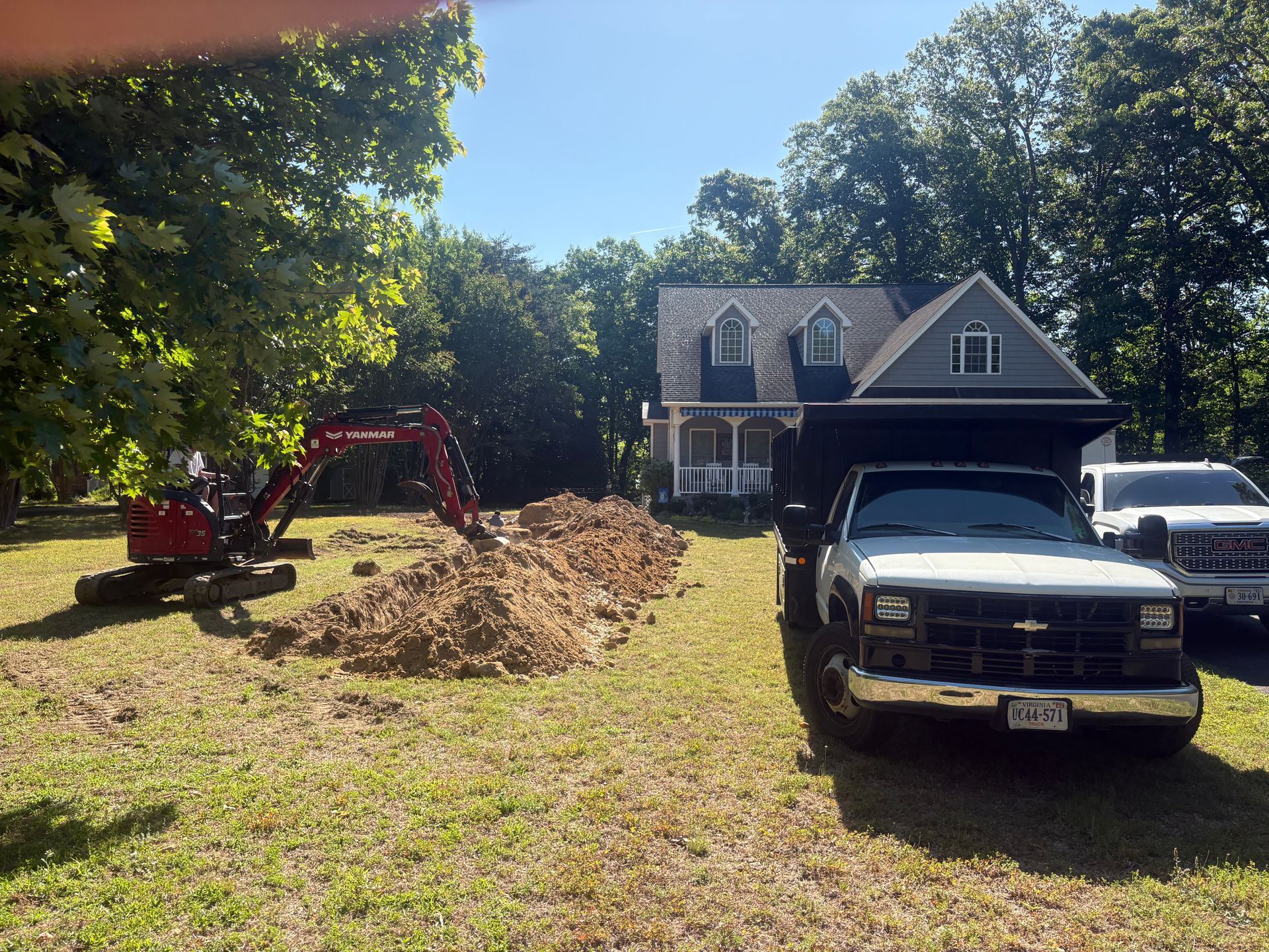 Suburban yard with a red excavator, large dirt pile, and two trucks in front of a house