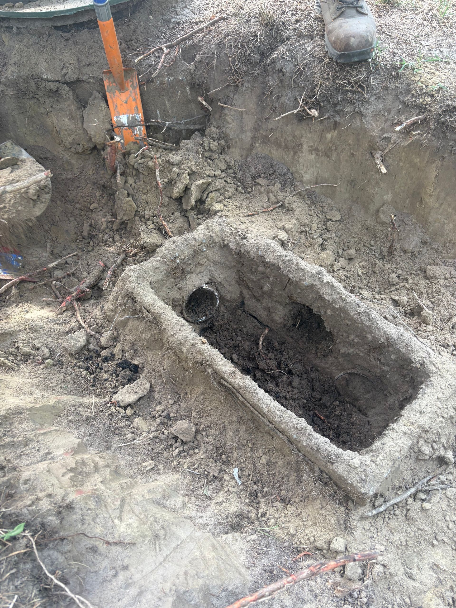 Excavation site with a muddy rectangular hole and an orange strap or tool at the edge.