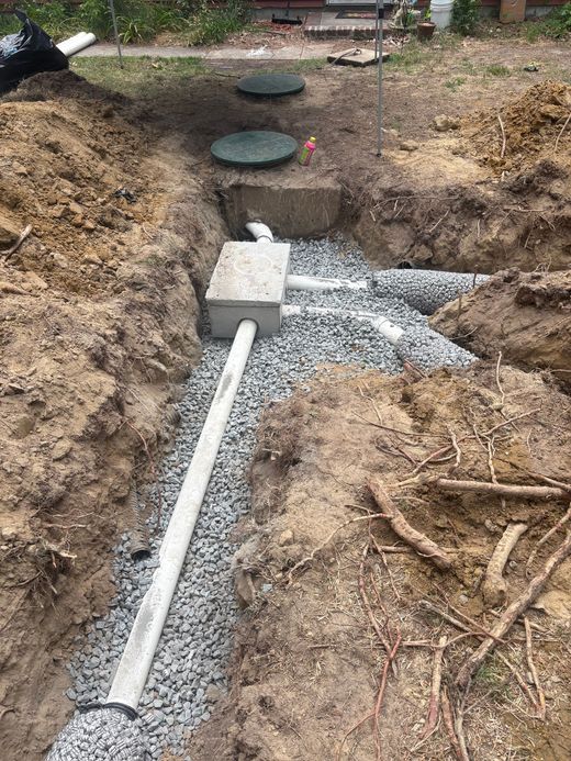 Trenched gravel drainage area with white pipe, concrete blocks, and a round green cover outdoors