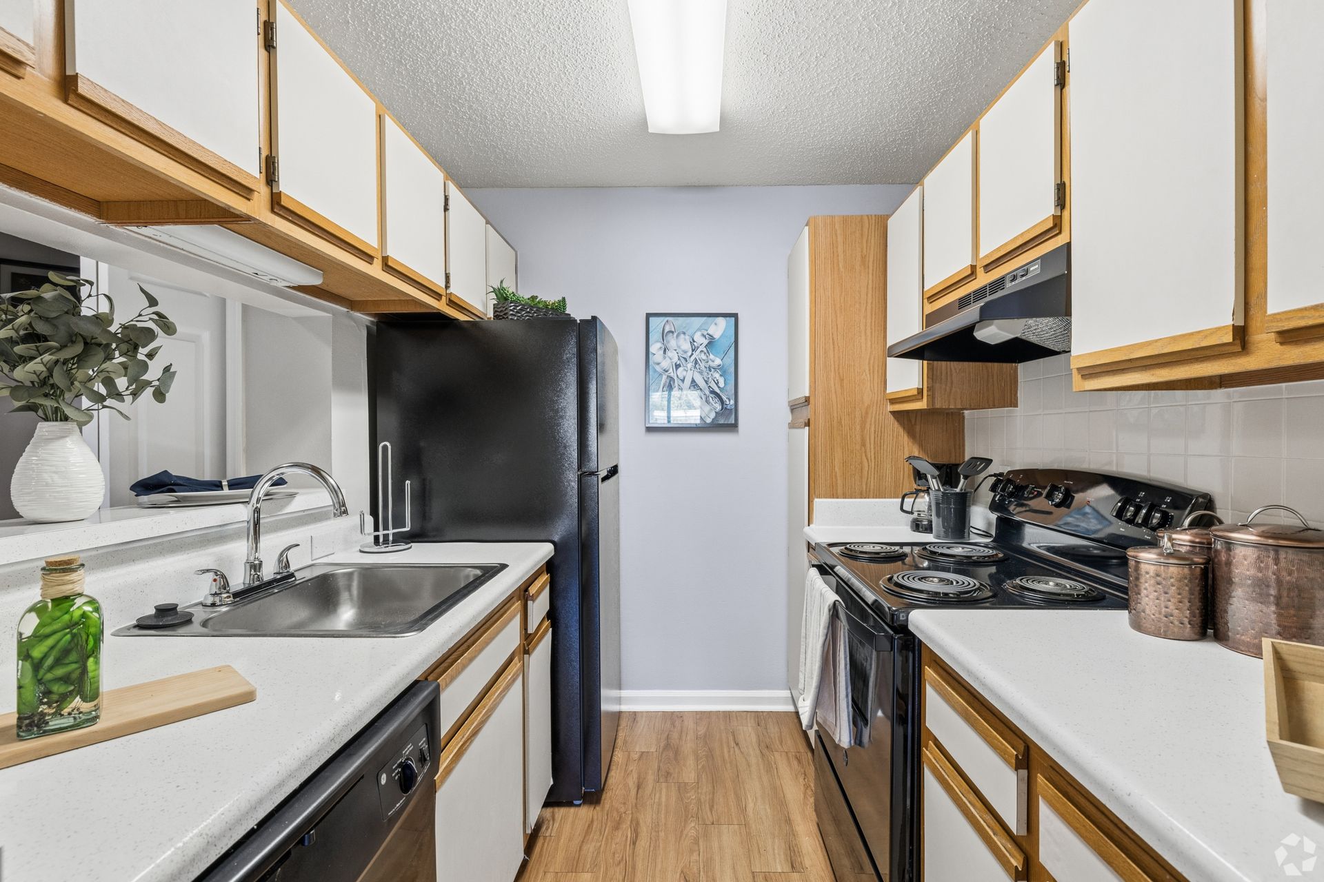 Narrow galley kitchen with white cabinets, wood accents, black appliances, and a window over the sink.