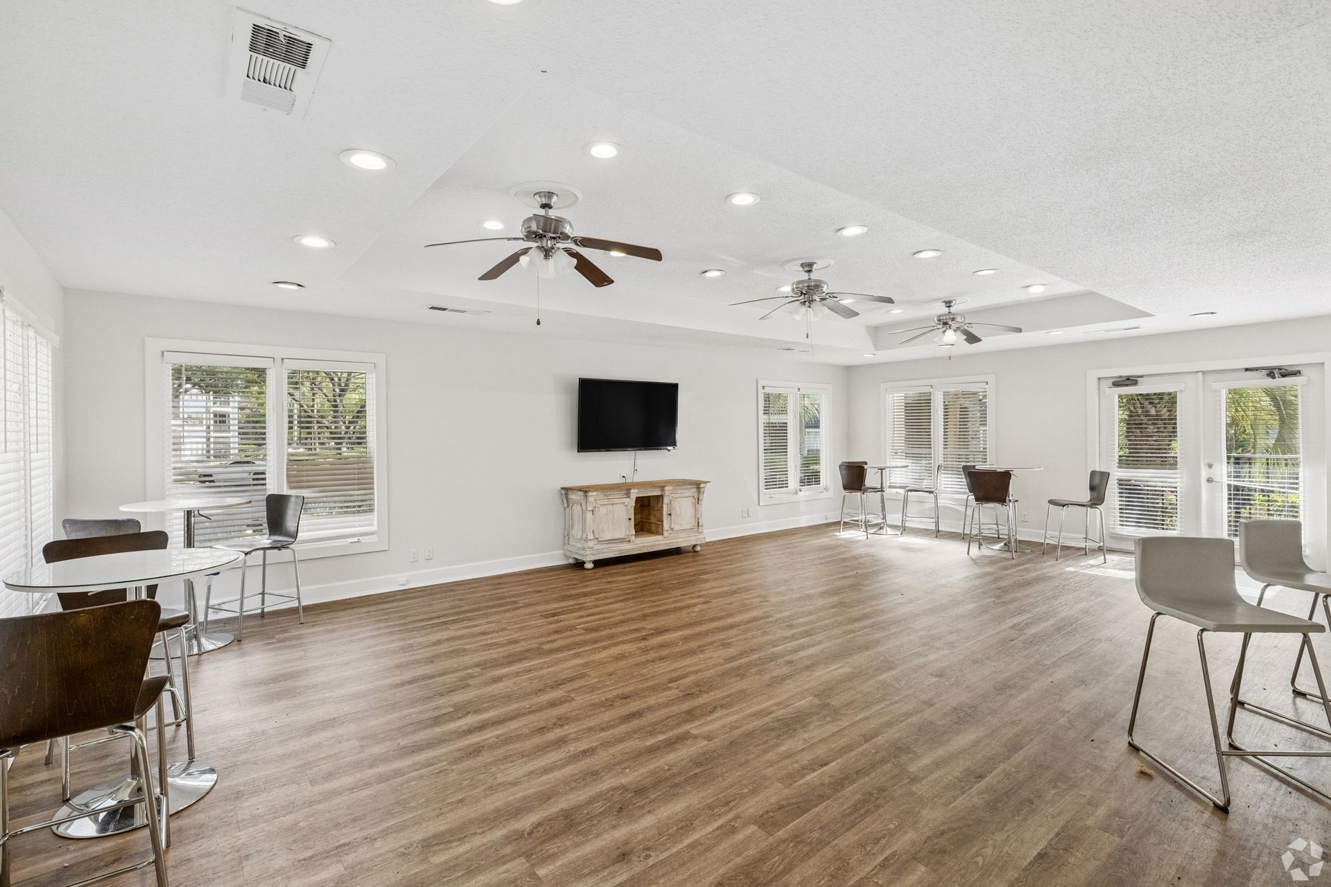 Bright empty community room with wood floors, ceiling fans, TV, and tables by large windows