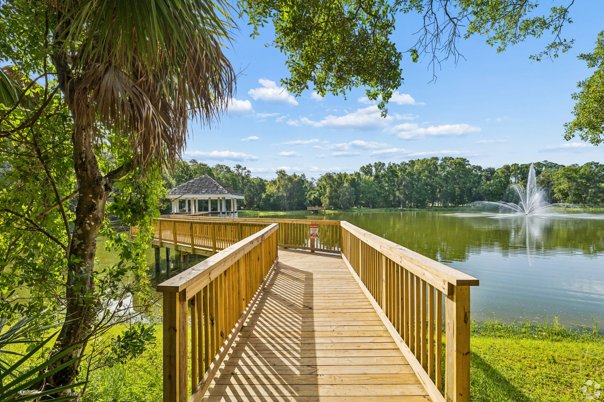 Wooden boardwalk leading to a lakeside pavilion and fountain, framed by palm trees and green grass