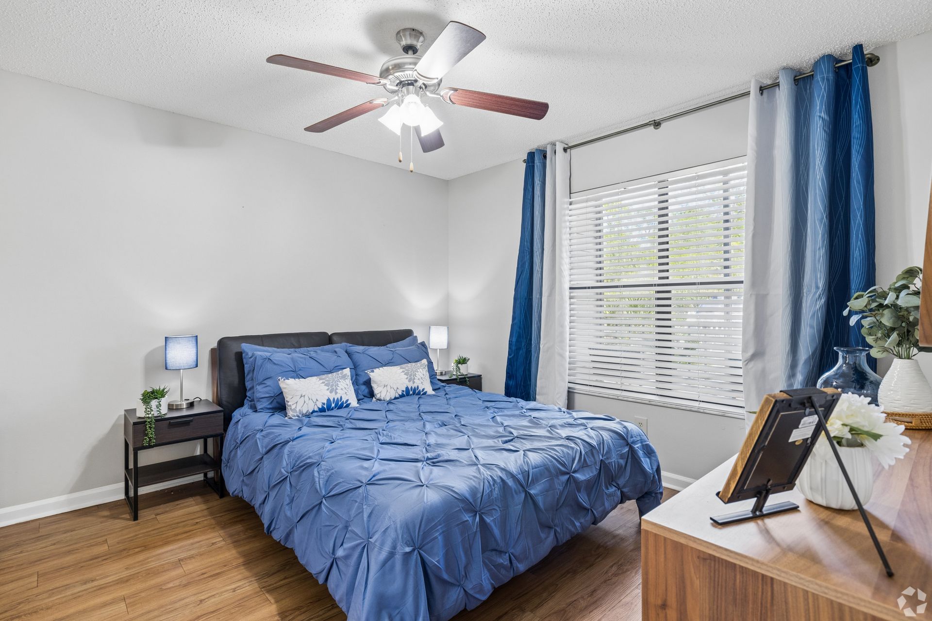 Bright bedroom with blue bedding, white walls, wooden floor, window, and ceiling fan.