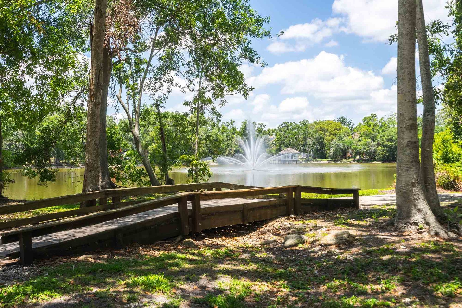 Park bench by a lake with a fountain, surrounded by trees and sunlight at Grande Oasis at Carrollwood apartments for rent in Tampa, Florida.