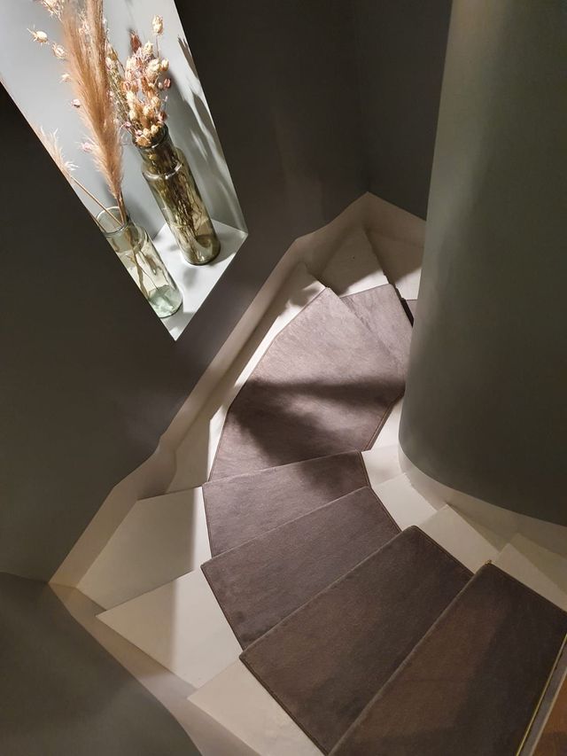 Vue plongeante d'un escalier courbe avec moquette foncée, contremarches blanches et murs gris. Une étagère décorative ornée de plantes séchées se trouve à gauche.