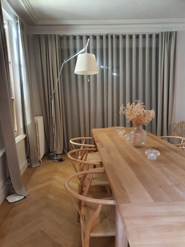 Salle à manger avec une longue table en bois avec des chaises, un lampadaire et des rideaux aux tons neutres.