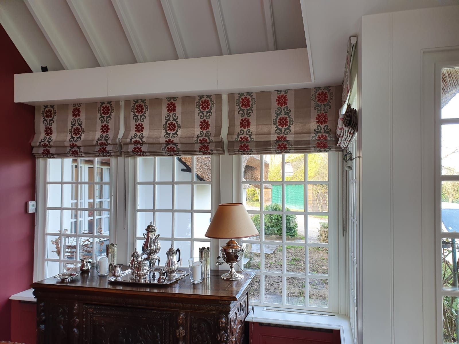 Une pièce avec une fenêtre ornée de stores romains à motifs floraux rouges, une armoire en bois et une lampe.