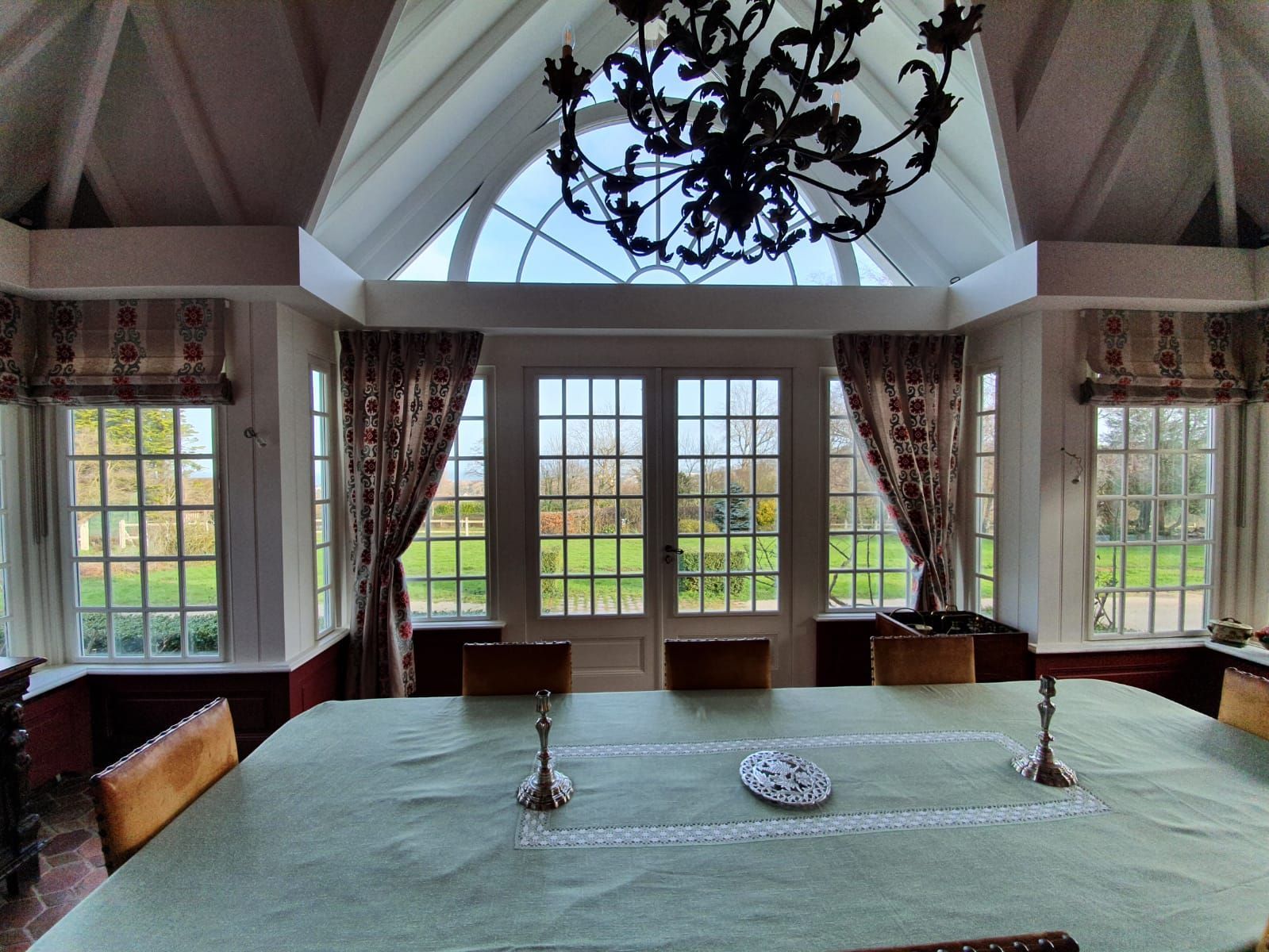 Une salle à manger ensoleillée avec une grande table dressée pour un repas, donnant sur une cour verdoyante à travers de grandes fenêtres et des portes-fenêtres.