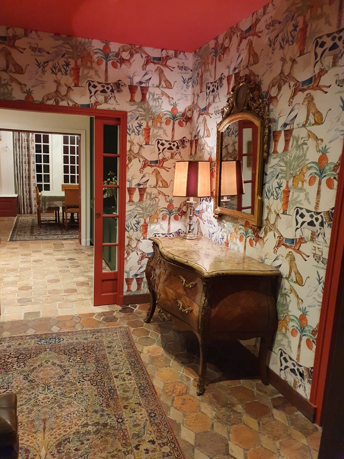 Une commode antique ornée avec un miroir dans un couloir avec du papier peint à motifs et une porte vers une autre pièce.