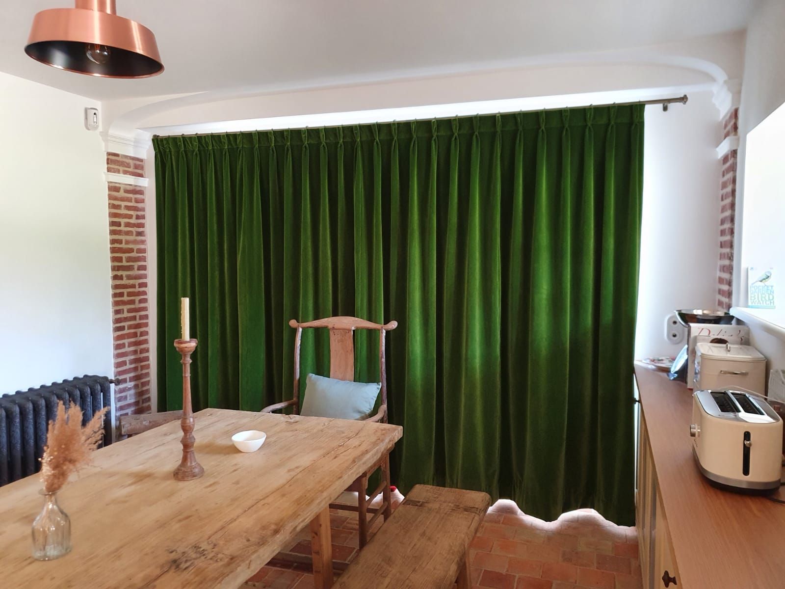 Une salle à manger avec des rideaux de velours vert vif couvrant une fenêtre. Au premier plan, une table et des chaises en bois, surmontées d'un luminaire en cuivre.