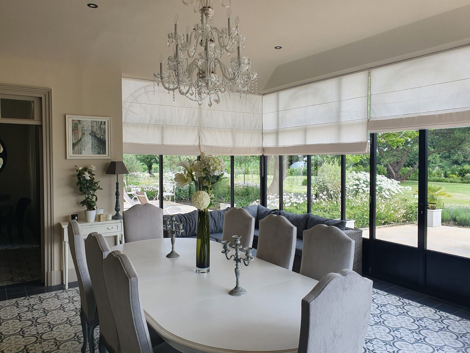 Salle à manger élégante avec une grande table blanche, un lustre orné et une vue sur un jardin.
