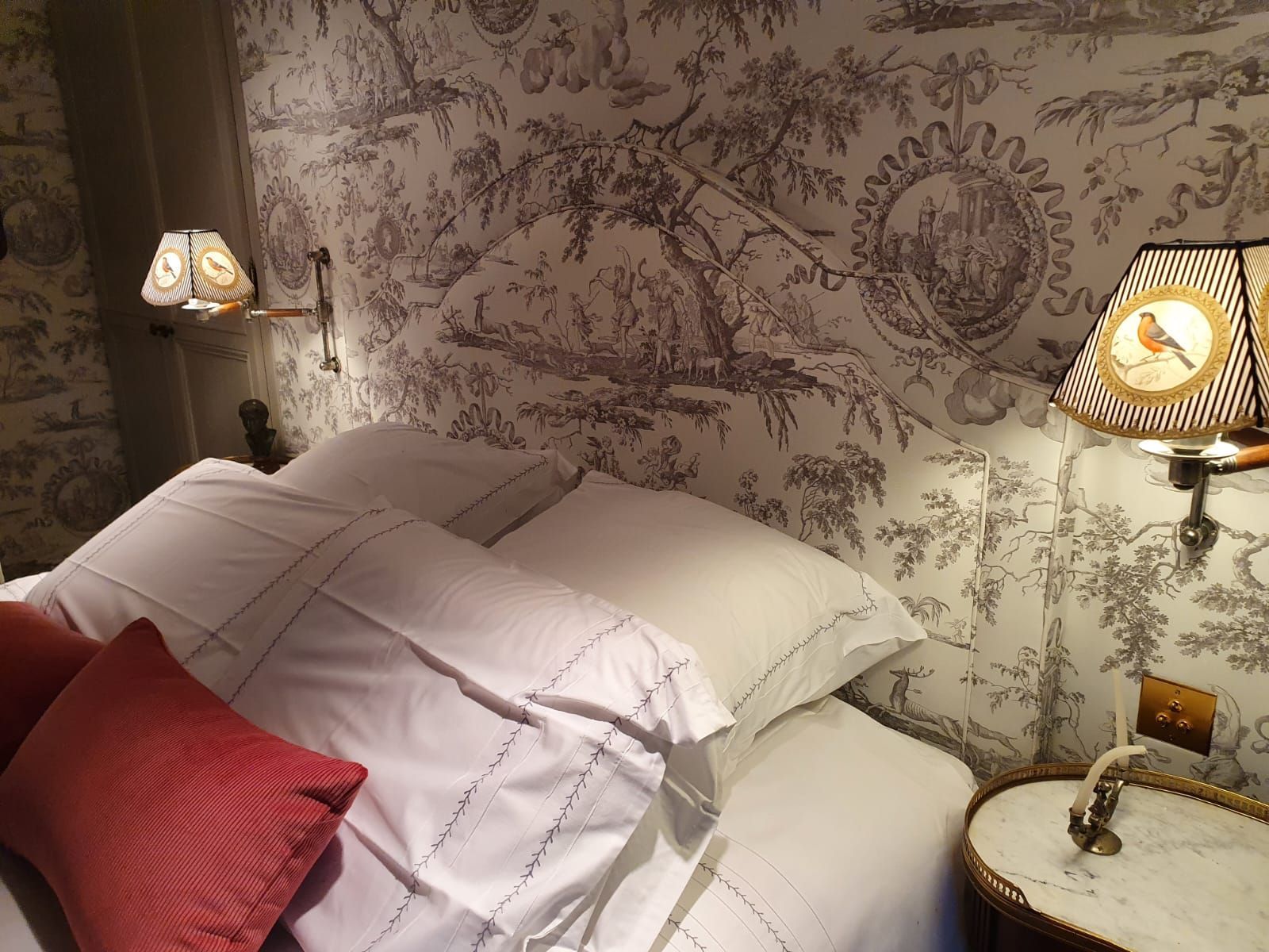 Un lit aux draps blancs et aux oreillers rouges, adossé à un mur recouvert de papier peint en toile de Jouy noir et blanc. Deux lampes aux abat-jour à motifs encadrent le lit.