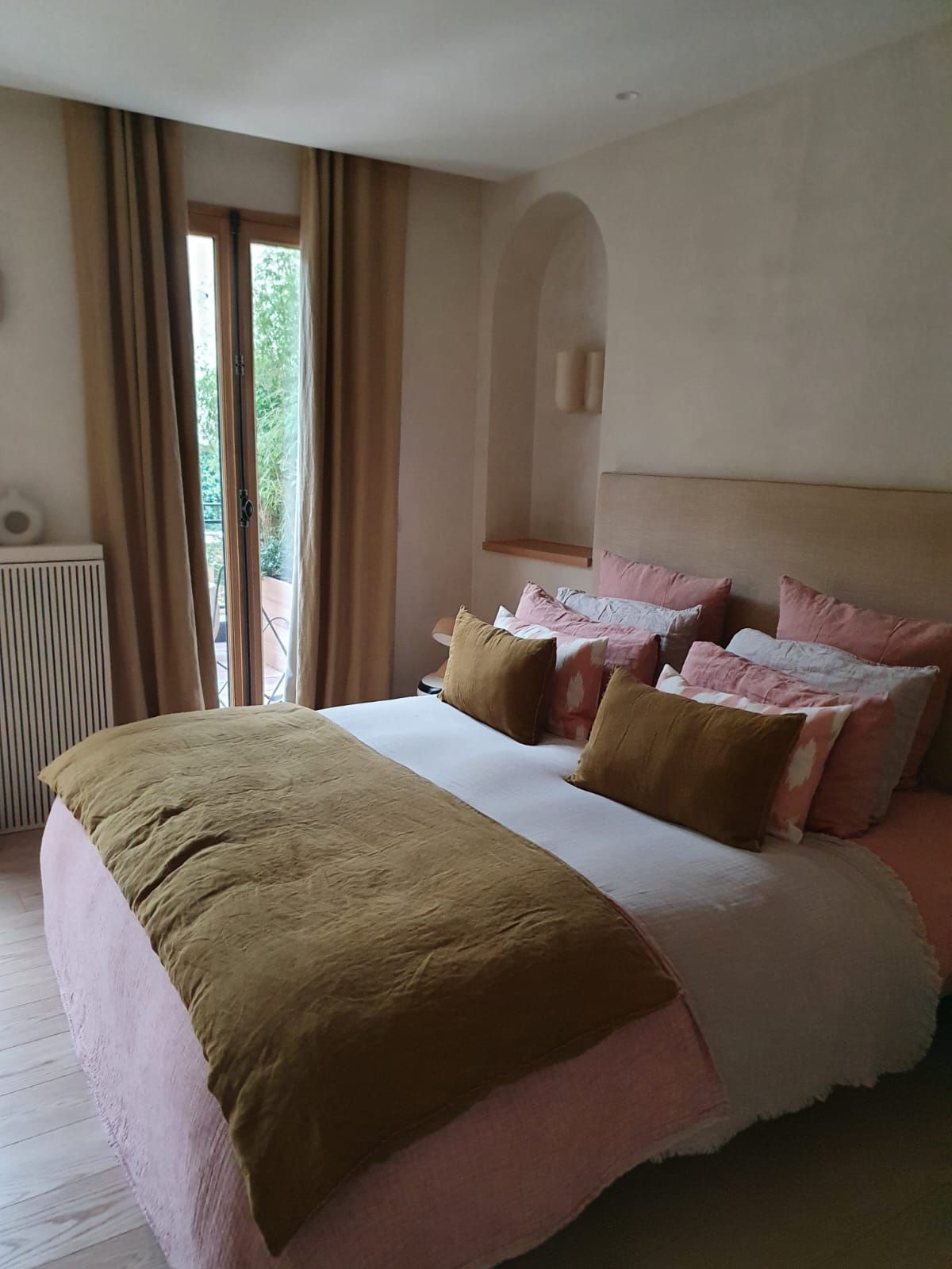 Une chambre avec un lit recouvert de draps roses et marron, près d'une fenêtre. Les murs sont clairs.