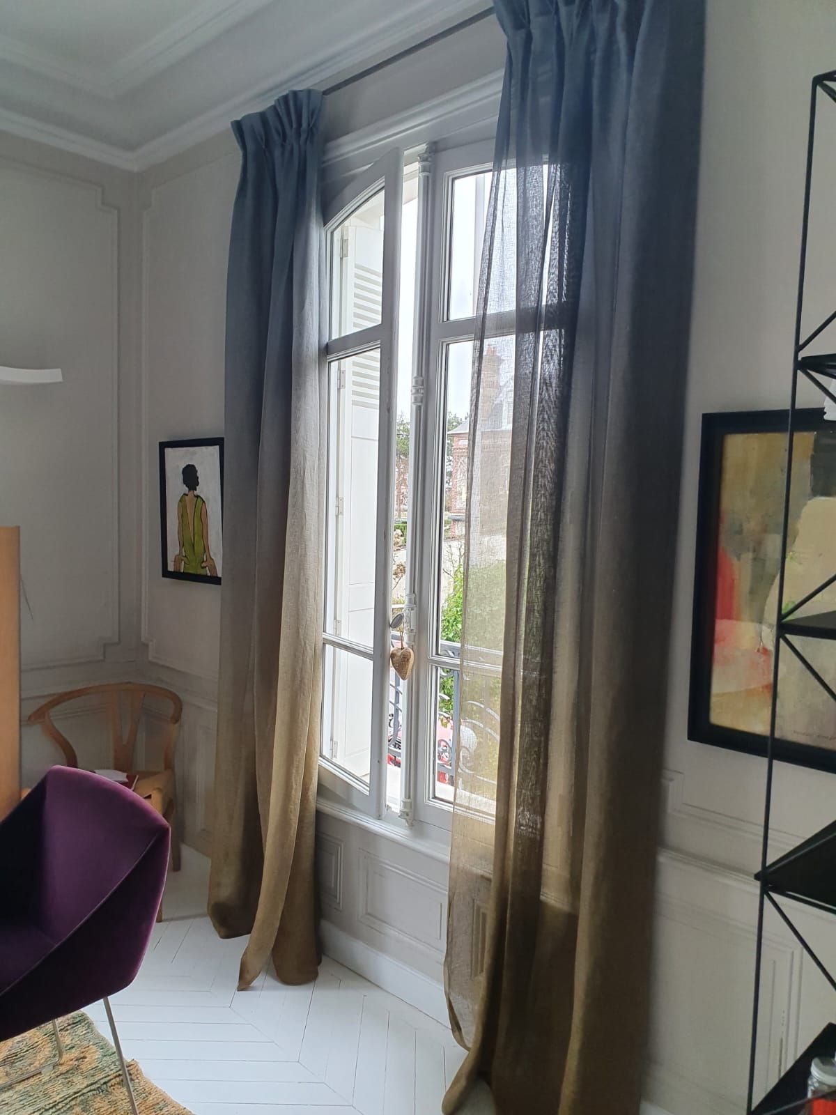 Une pièce avec une grande fenêtre ouverte encadrée de rideaux dégradés bleu-brun. On y voit une chaise violette et des œuvres d'art.