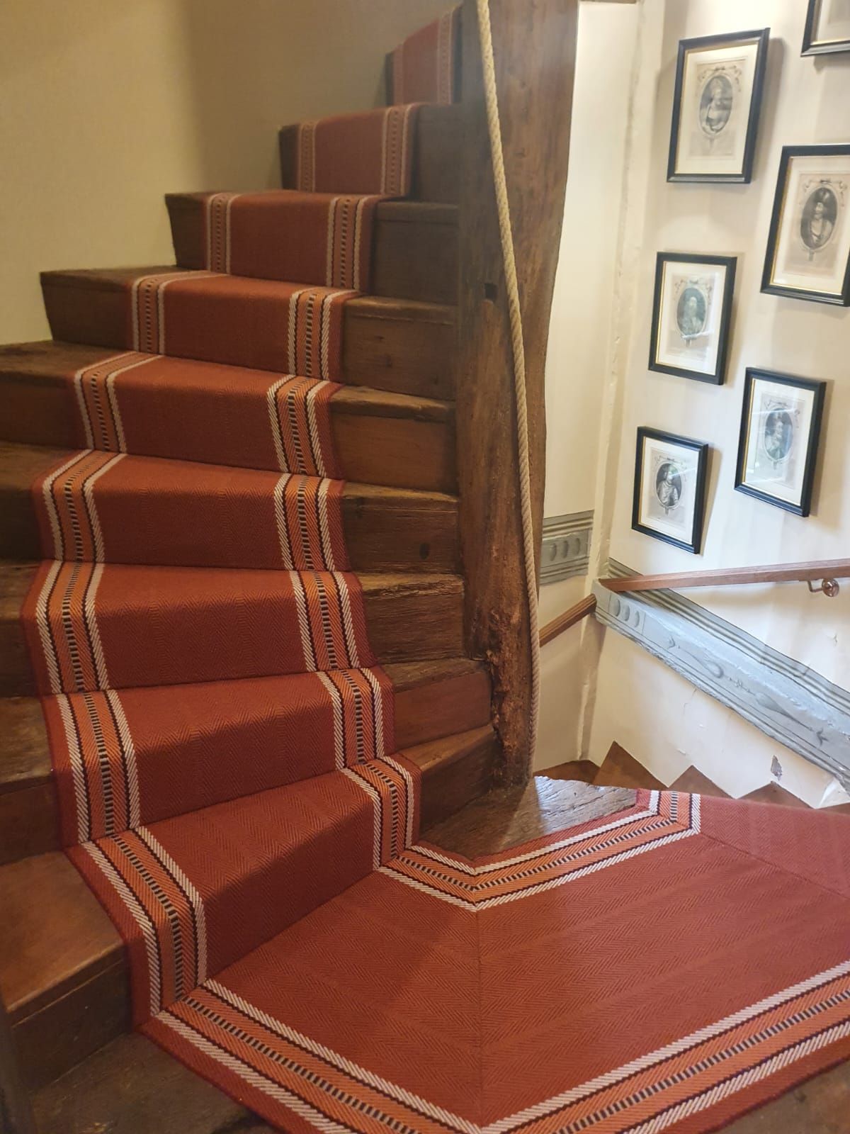 Escalier en bois avec tapis rouge et moulures décoratives. Des portraits sont accrochés au mur crème à droite.